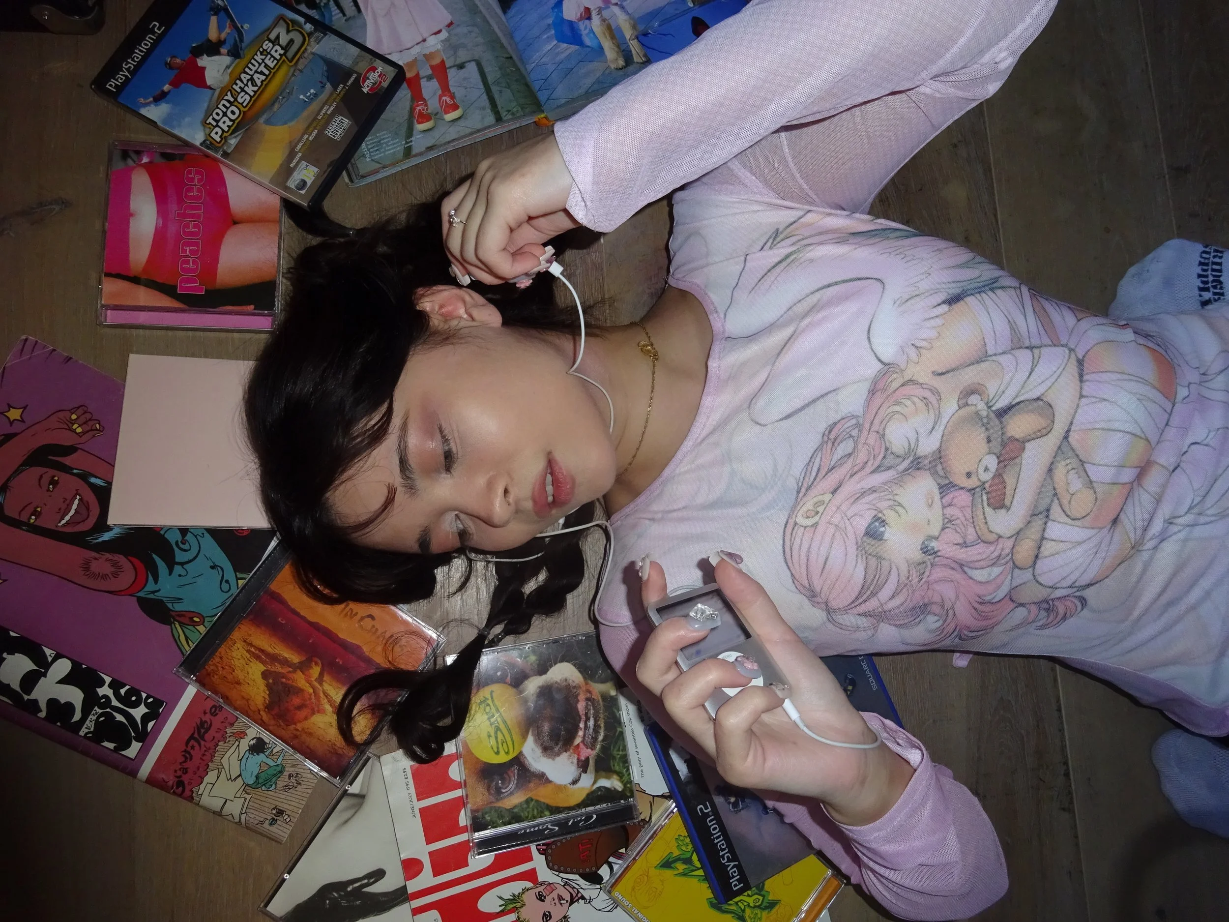 A woman lying on the floor with dark hair, wearing pink pajamas with cartoon characters, holding a gaming controller, surrounded by various CDs, DVDs, and magazines. The floor is wooden, and there are posters and artwork on the wall.