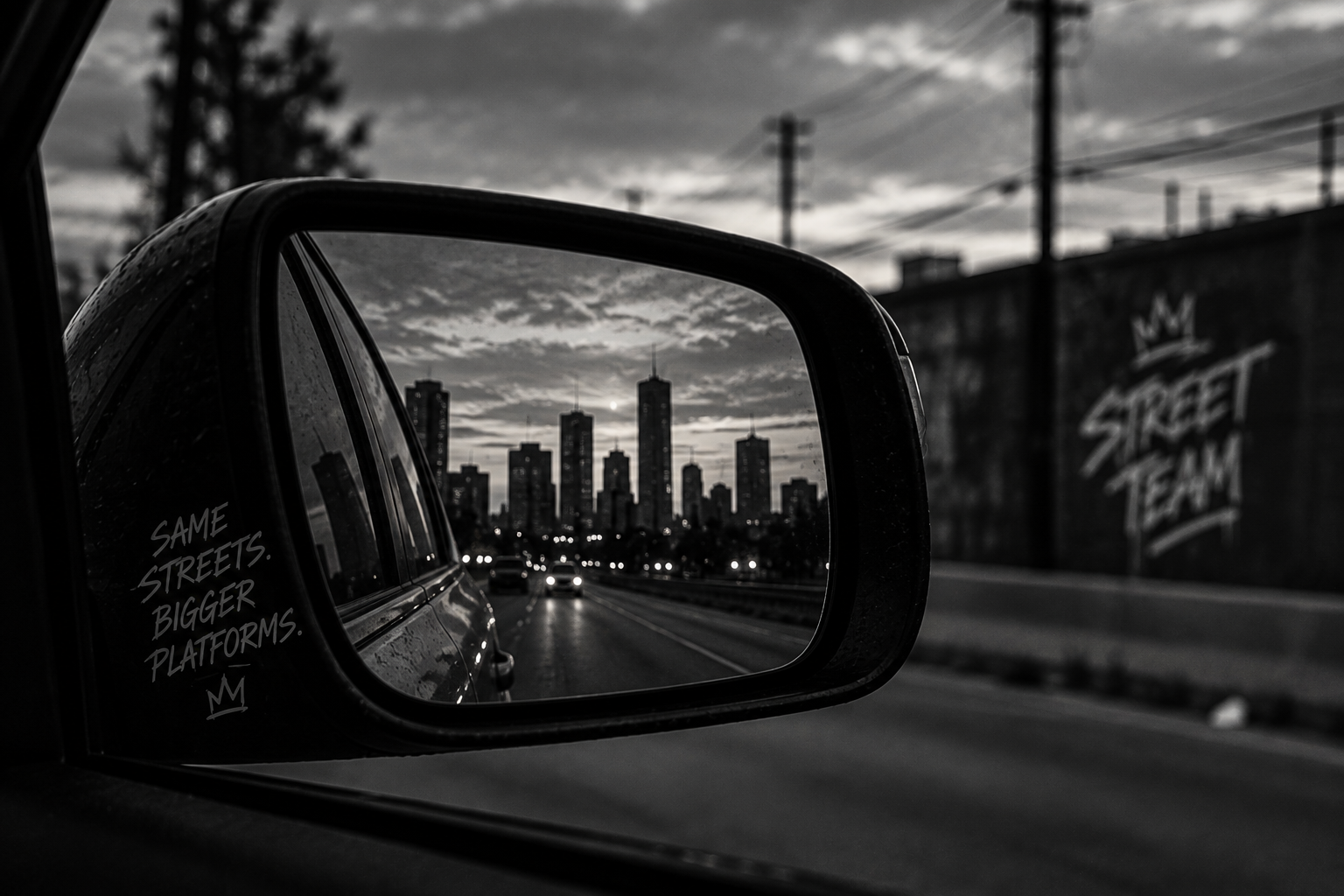 Black and white photo of a car's side mirror reflecting a city skyline with tall buildings and a cloudy sky. The mirror has a handwritten message: 'SAME STREETS. BIGGER PLATFORMS.'