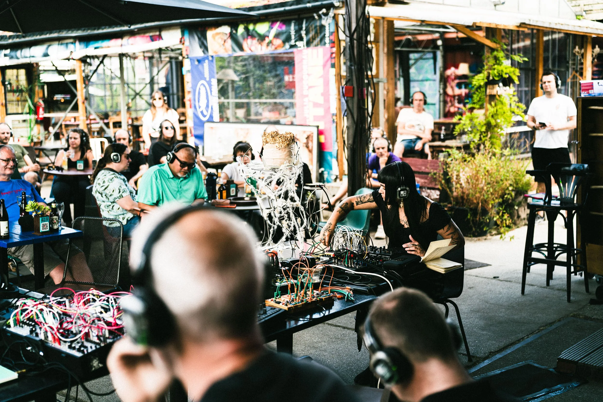 A crowd of people outdoors attending a live music event with a DJ and electronic equipment on a table, some people wearing headphones and sunglasses, surrounded by plants and banners.