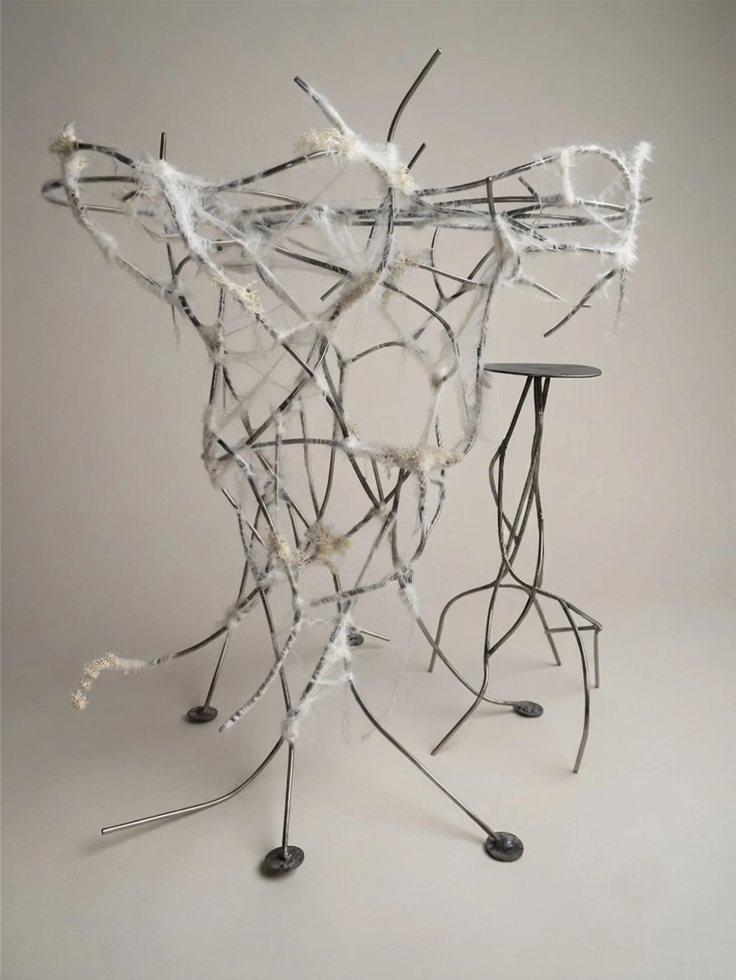 Abstract metal sculpture resembling mycelium with thin metal rods, some wrapped with white fabric or fibers, arranged on a plain background.