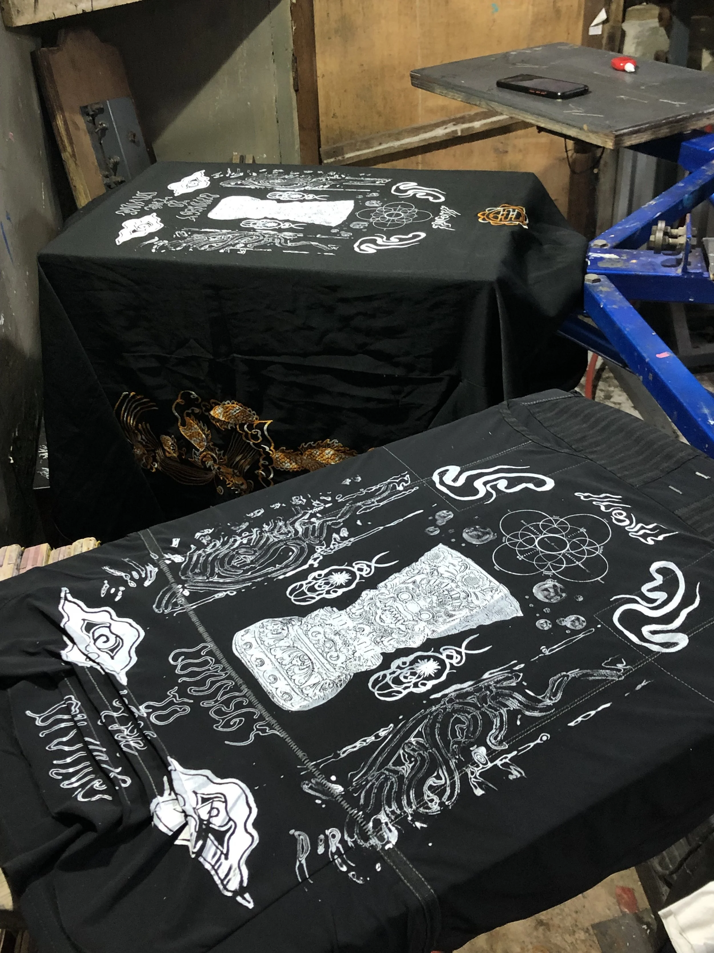 Black cloth with white intricate designs and drawings spread on a screen-printing table.
