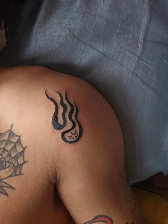 Tattoo of a flame with a face on shoulder, spider web tattoo on chest, and other tattoos on arm.