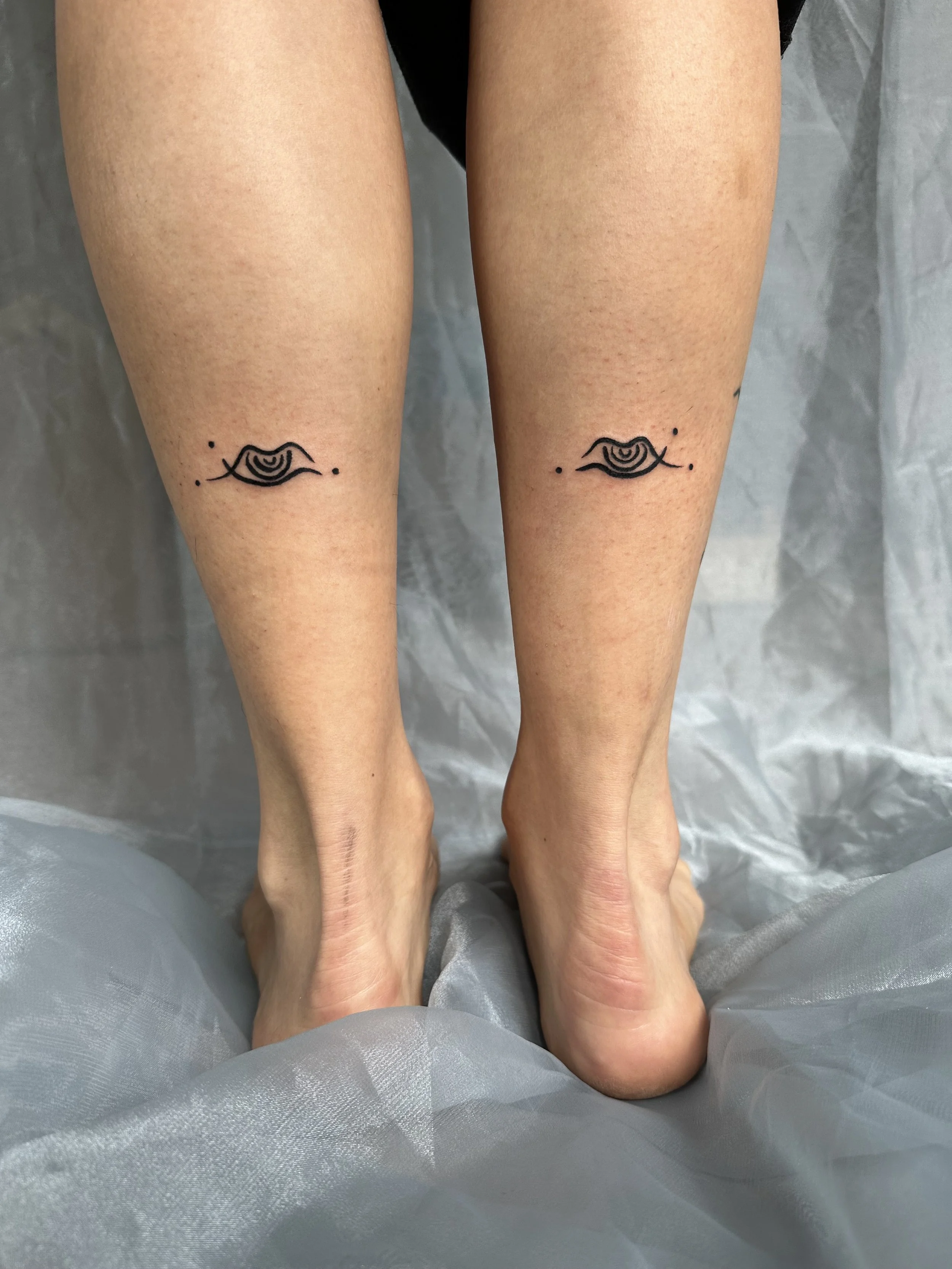 Close-up of two person's legs with matching eye tattoos just below the knees, standing on a gray, textured surface.