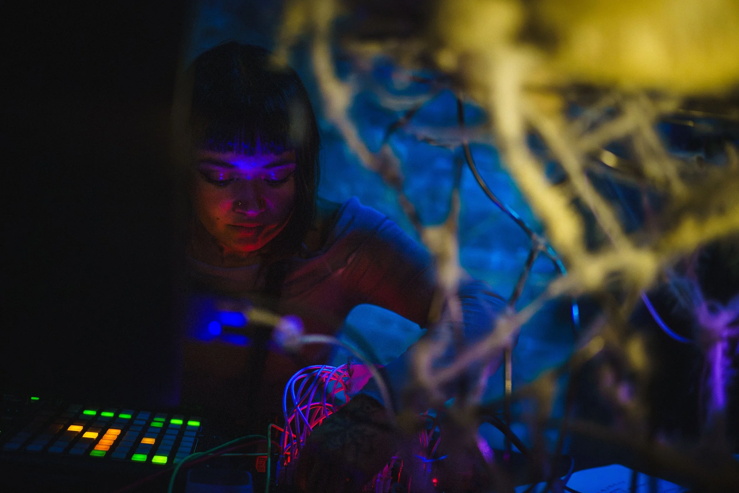 A person  illuminated by colorful LED lights, sitting at a table with electronic music equipment, surrounded by glowing wires and a colorful grid pad in a dark setting.