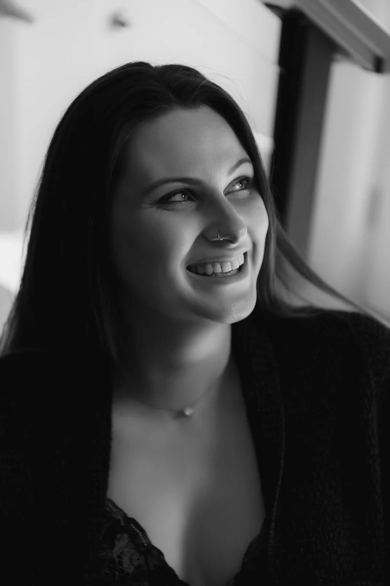 A woman with dark hair smiling and looking to her left, wearing a nose ring, a necklace, and a dark top in a black and white photo.