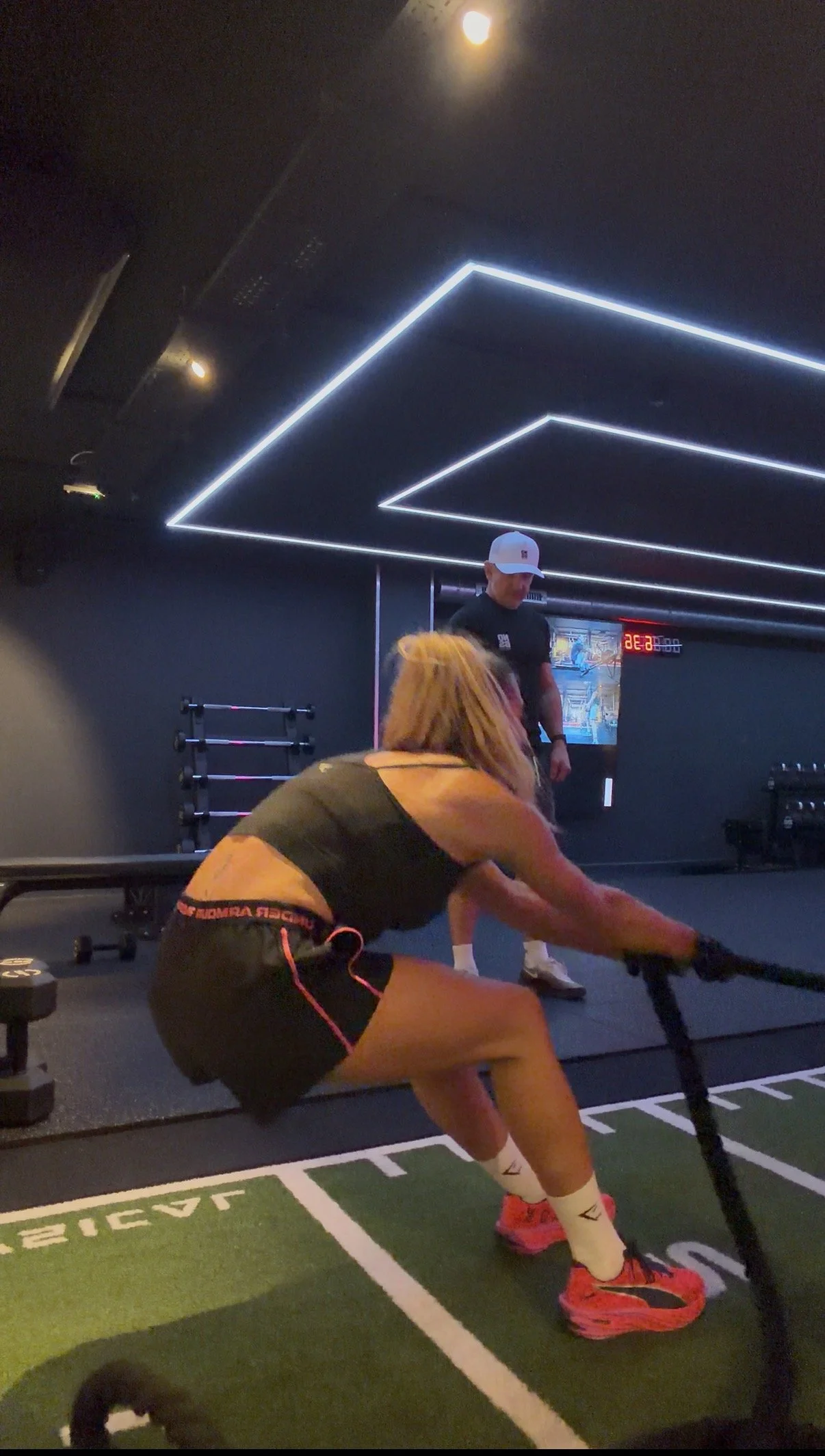 A woman in workout clothes doing a rowing exercise with a trainer supervising in a gym with modern lighting.