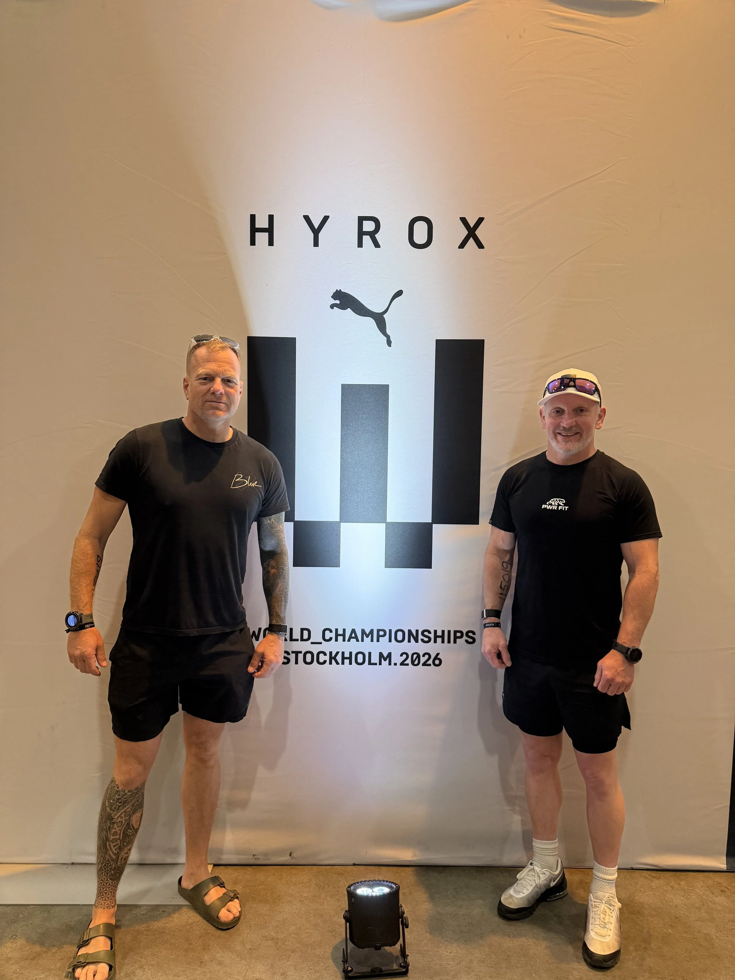 Two men standing in front of a backdrop that reads 'HYROX' with a Puma logo, and 'WORLD CHAMPIONSHIPS STOCKHOLM 2026.' The man on the left is wearing black shorts, a black t-shirt, sandals, and sports watches, with tattoos on his arms and a patterned tattoo on his leg. The man on the right is wearing black shorts, a black t-shirt, white sneakers, a white cap with sunglasses on it, and a sports watch, with tattoos on his arms.