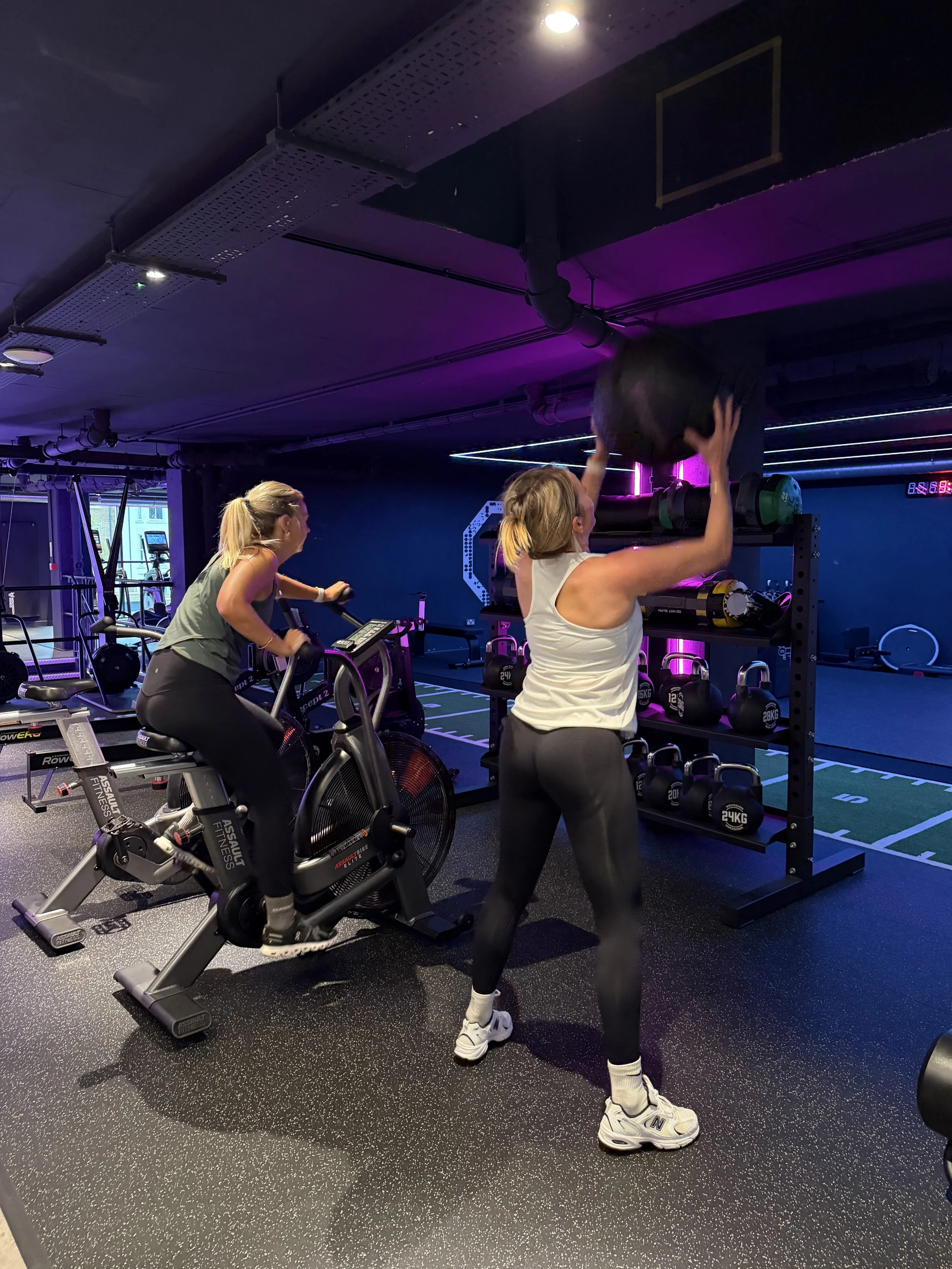 Two women working out at a modern gym with black and neon purple lighting; one on a stationary bike, the other lifting a weight on a rack.