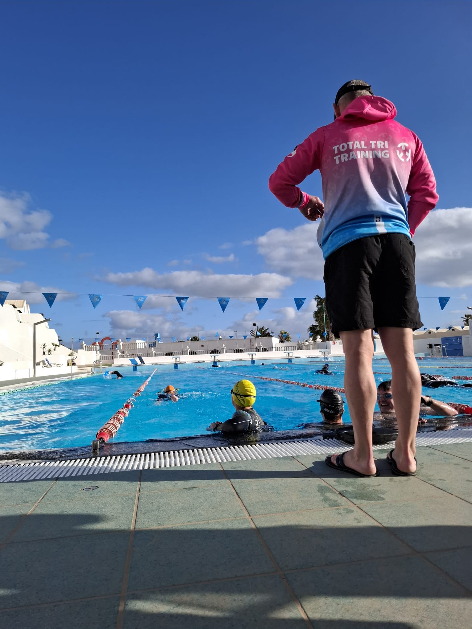 Swimmers in a pool with a coach or lifeguard standing at the edge, overlooking the activity, under a blue sky with some clouds. Blue triangular flags stretch across the background.
