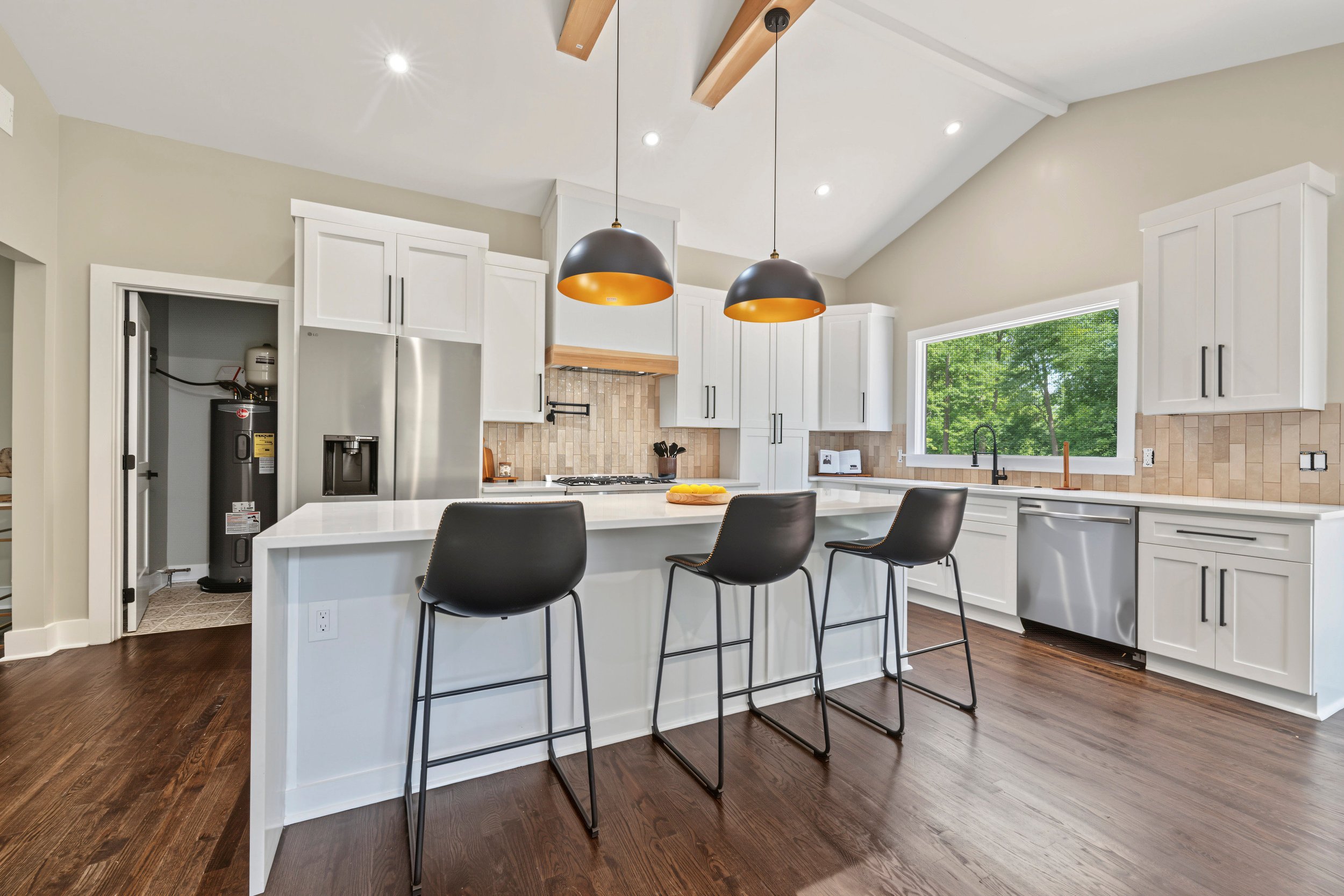 Modern white kitchen with dark wood flooring, large window showing green trees, island with black chairs, pendant lights, and built-in appliances.