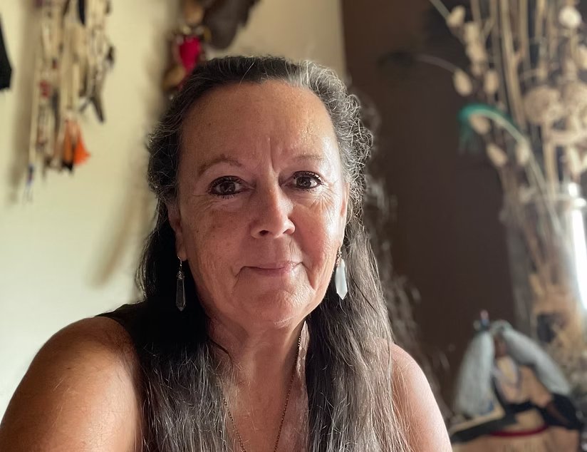 An older woman with long gray hair, wearing earrings, smiling in a room with artwork and decorations on the wall behind her.