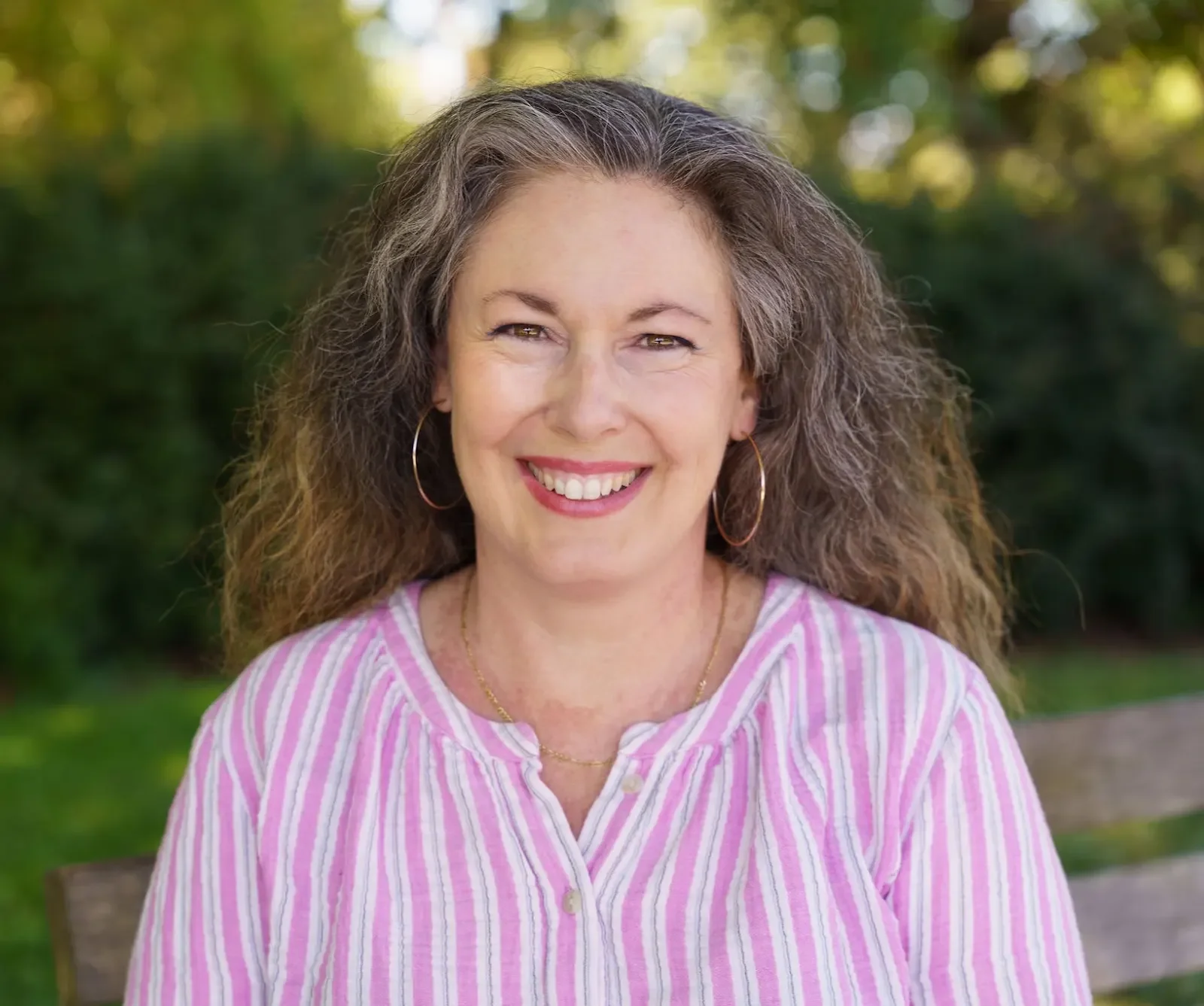 A woman with curly brown hair smiling outdoors, wearing gold hoop earrings and a pink and white striped blouse.