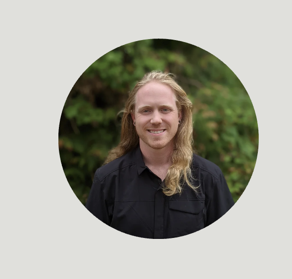 Headshot of a smiling man with long blond hair, wearing a black shirt, outdoors with greenery in the background.