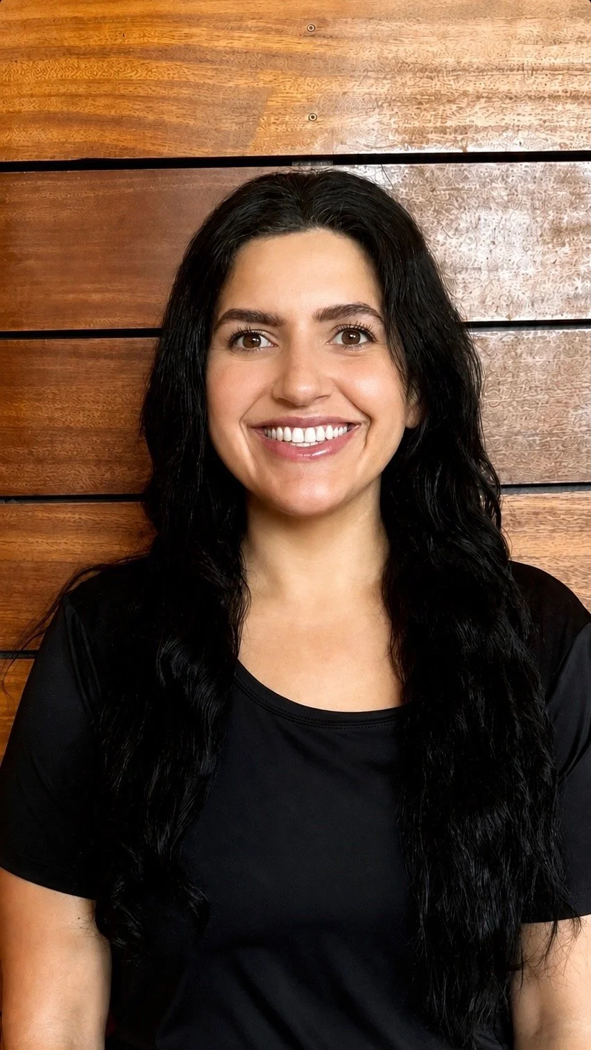 A woman with long black wavy hair, smiling, wearing a black shirt, standing in front of a wooden wall.