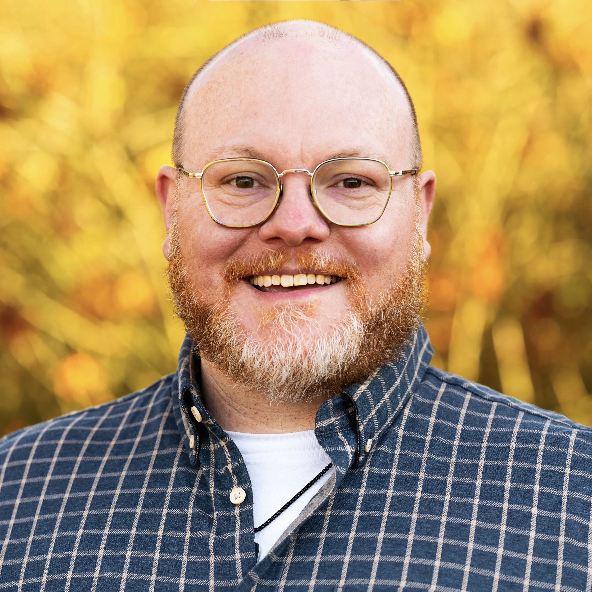 Close-up of a smiling man with glasses, a bald head, reddish beard, wearing a black and white plaid shirt, against a blurred yellow and orange background.