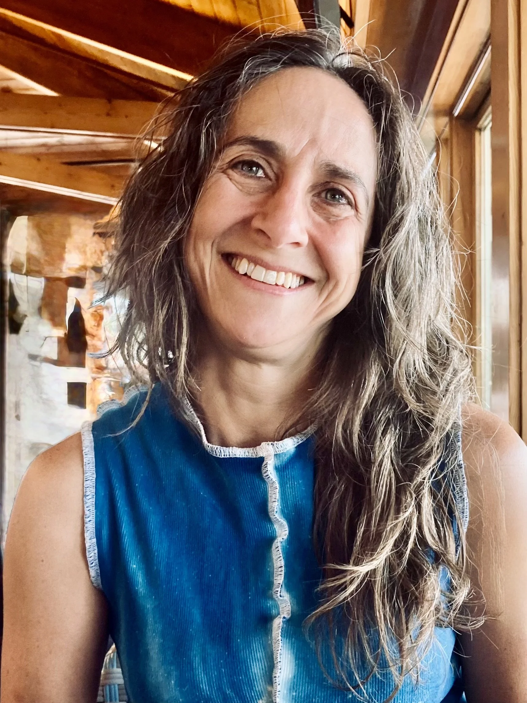 Close-up of a woman with long, curly grayish-brown hair smiling, wearing a blue sleeveless top with white trim, sitting indoors near wood-paneled walls and large windows.