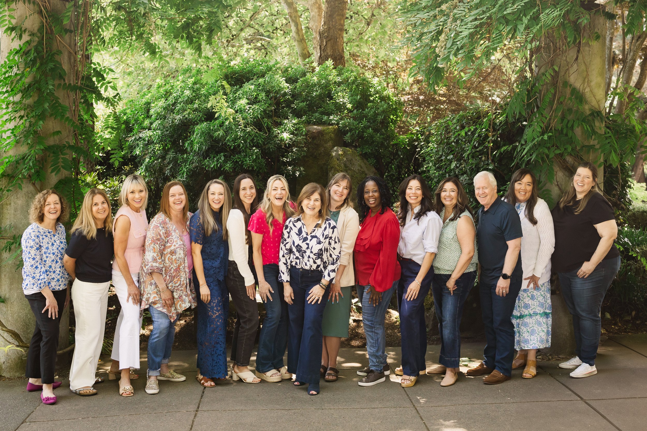 A diverse group of 16 women and 2 men standing outdoors in front of lush green trees and foliage, smiling and posing for a photo.
