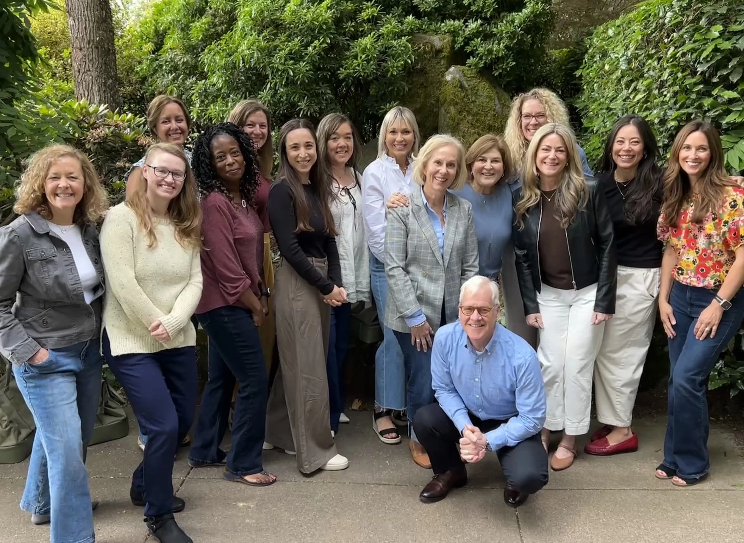 A group of fifteen women and one man posing outdoors in front of lush green bushes and trees. The women are smiling and dressed in casual and semi-formal attire, with some wearing glasses. The man, kneeling in front, is wearing a light blue dress shirt and dark pants.