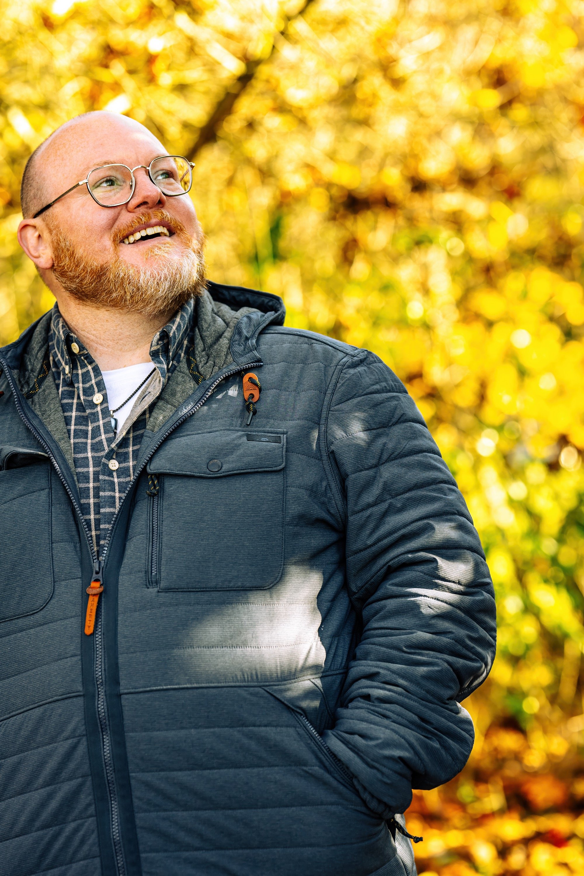 A smiling man with glasses and a beard looking upwards outdoors in fall, wearing a dark gray jacket and a plaid shirt, with yellow and orange autumn leaves in the background.