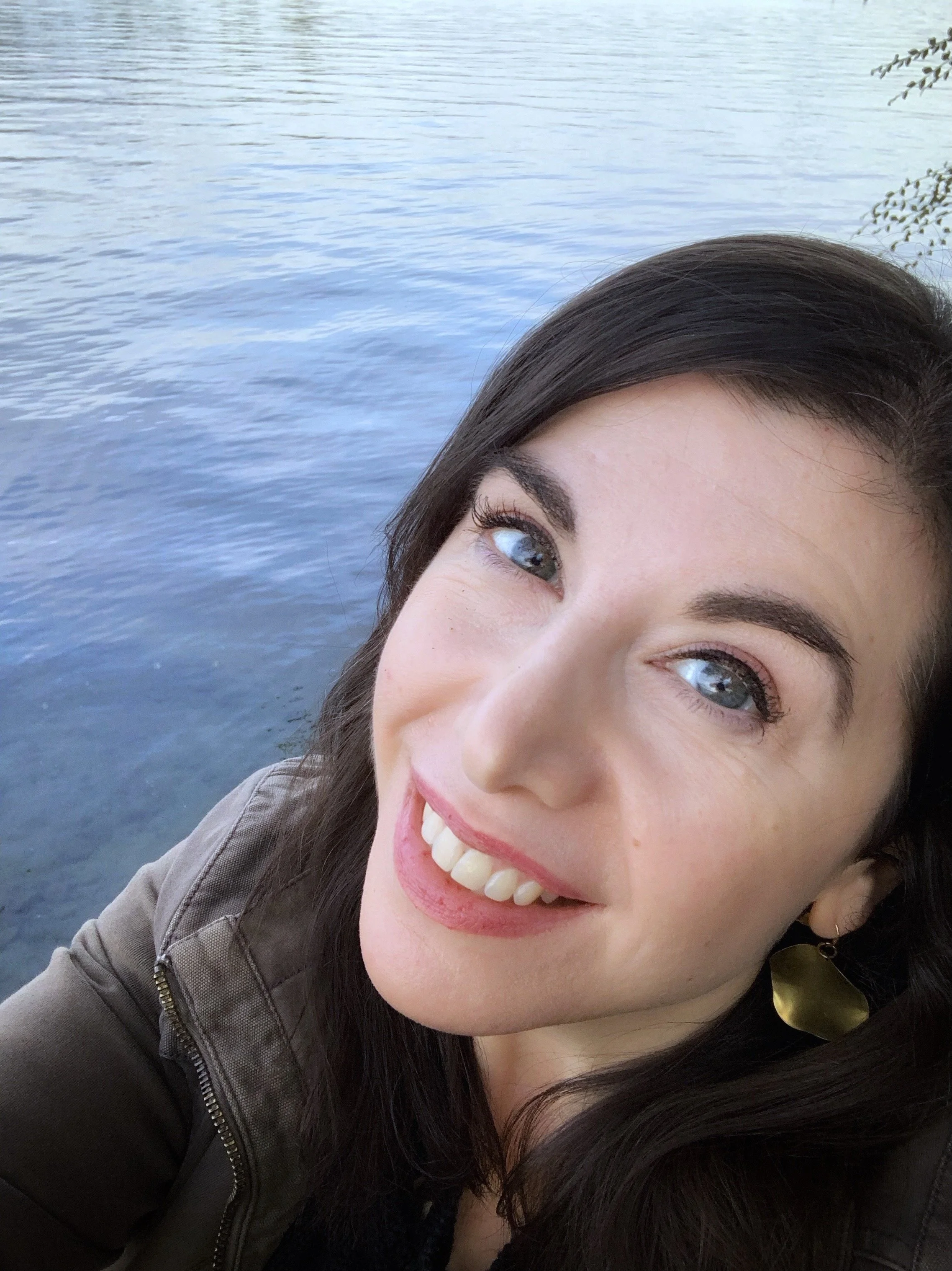 A close-up selfie of a woman with dark hair and blue eyes, smiling and showing her teeth, with a body of water in the background.