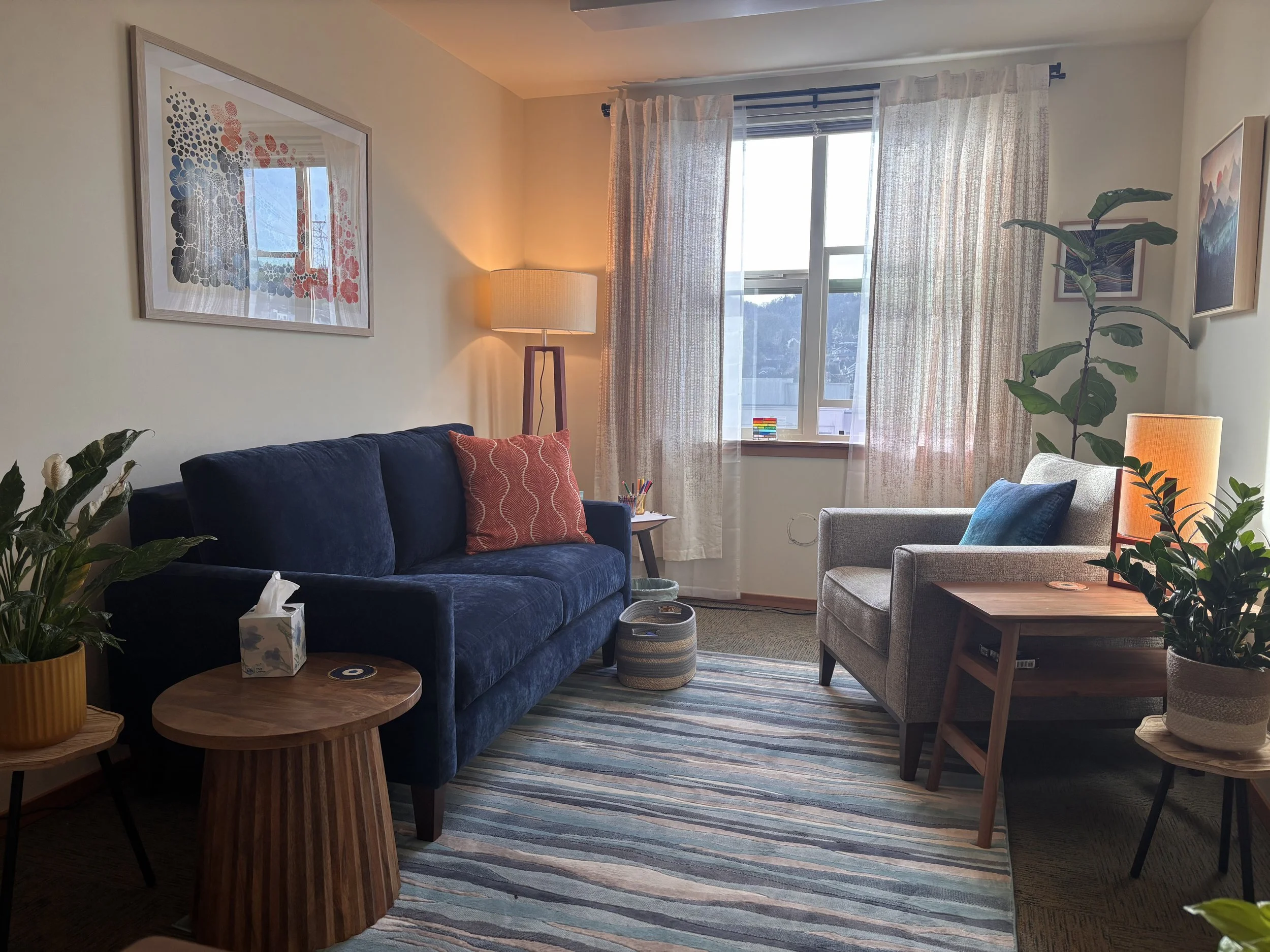 A living room with a navy-blue sofa, a wooden coffee table, and a gray armchair. There are colorful pillows on the sofa and a woven basket next to the armchair. A window with sheer curtains allows natural light, and framed artwork decorates the light-colored walls. Indoor plants are placed around the room, and a standing lamp provides additional lighting.