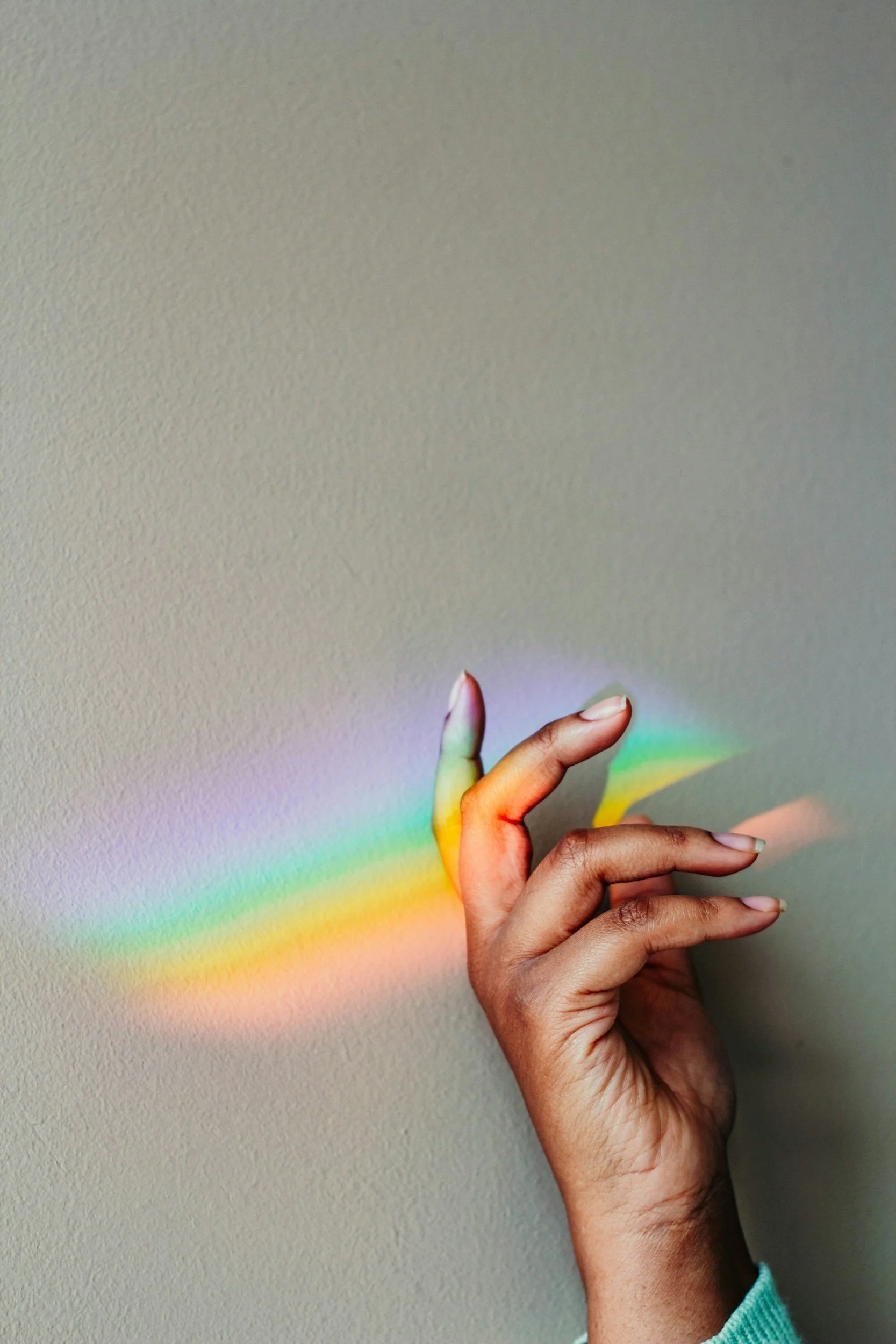 A hand with dark skin is holding a rainbow-colored light projection on a plain wall.