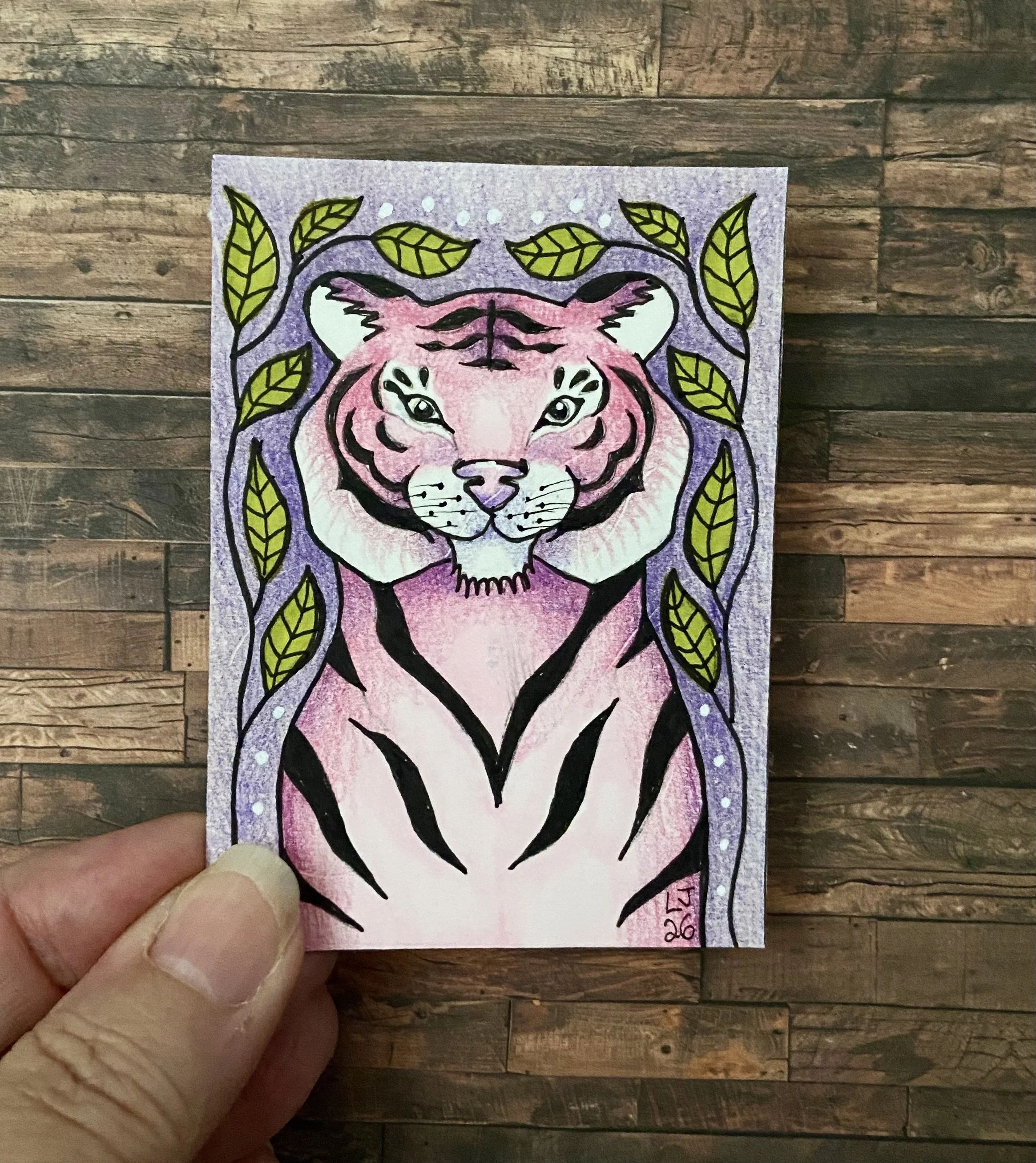 Colorful illustration of a tiger face surrounded by green leaves on purple background, with white dots, on a paper held between thumb and index finger. Wall behind wooden panels.