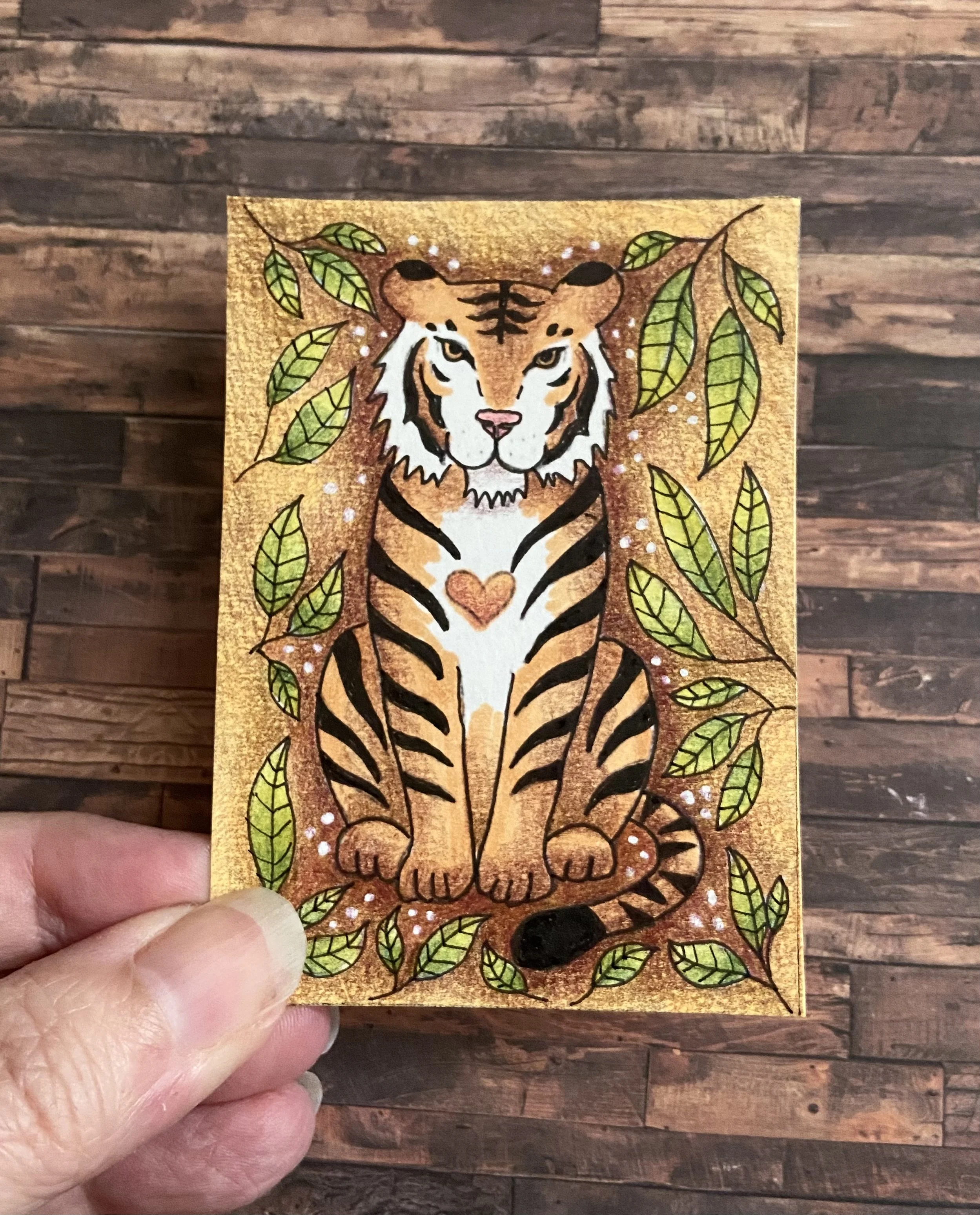 A colorful illustration of a tiger surrounded by green leaves and small white dots on a glittery gold background, held by a person's hand with a wooden floor in the background.