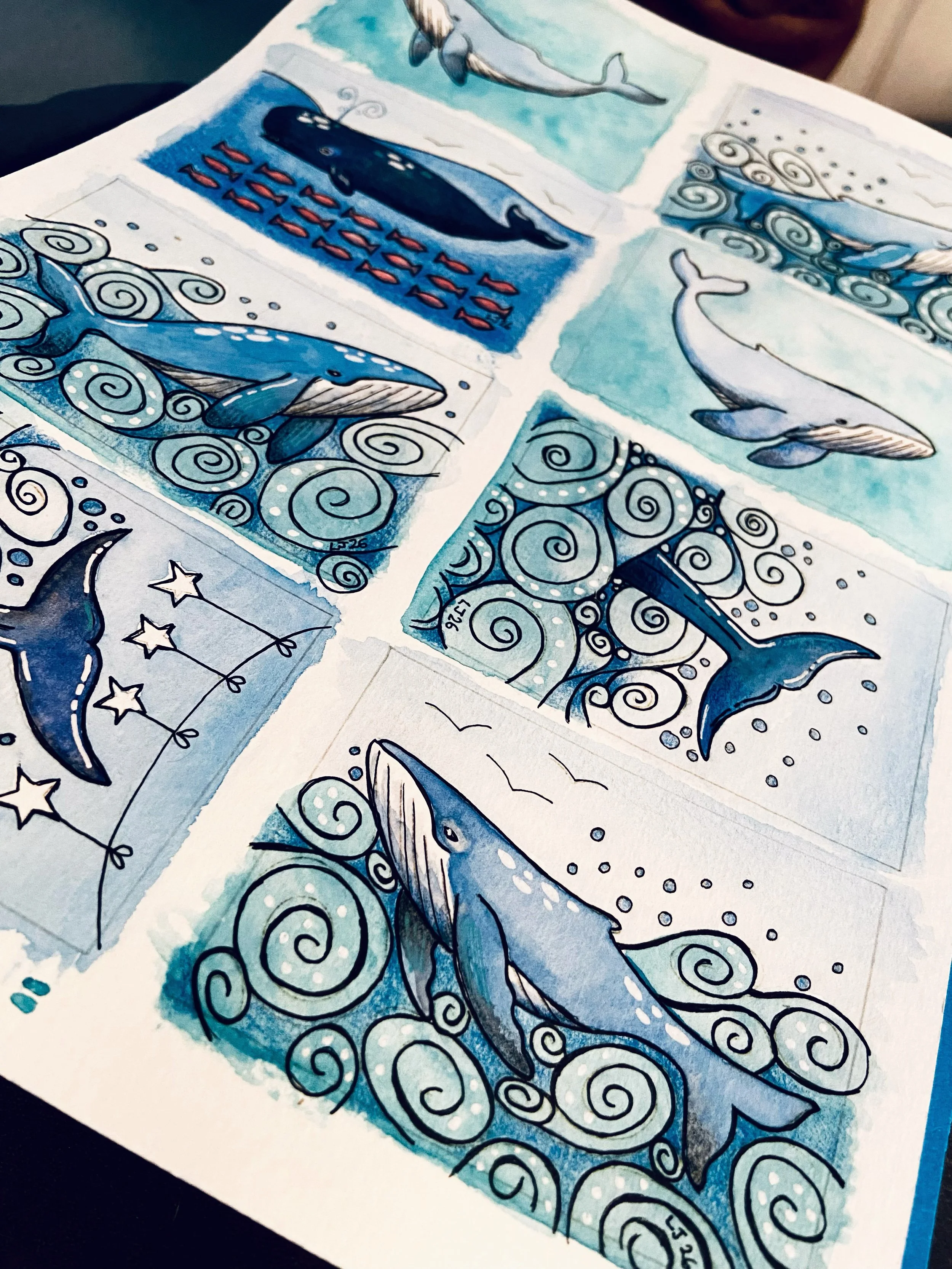 A close up of an A4 sheet of paper with eight drawings of whales in watercolour and colour pencil