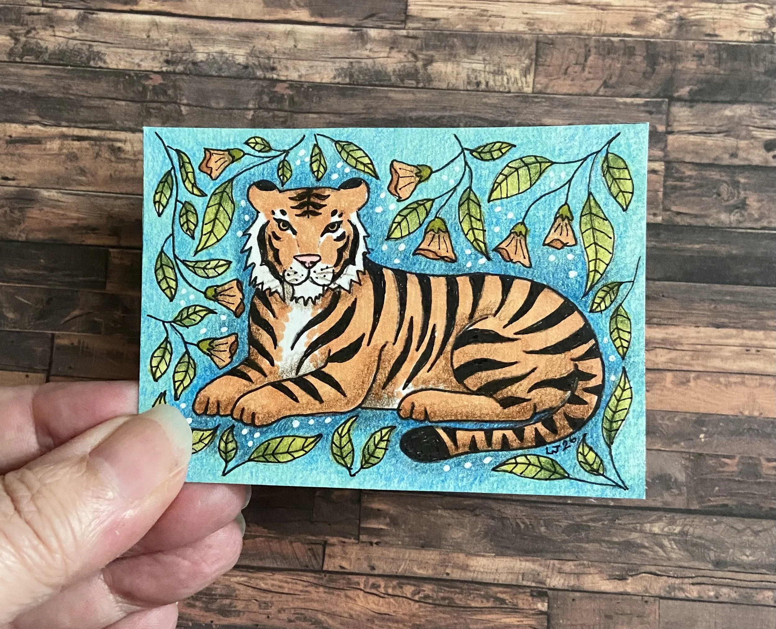 A hand holding a colorful drawing of a tiger lying amidst green leafy branches with orange flowers, on a blue background.