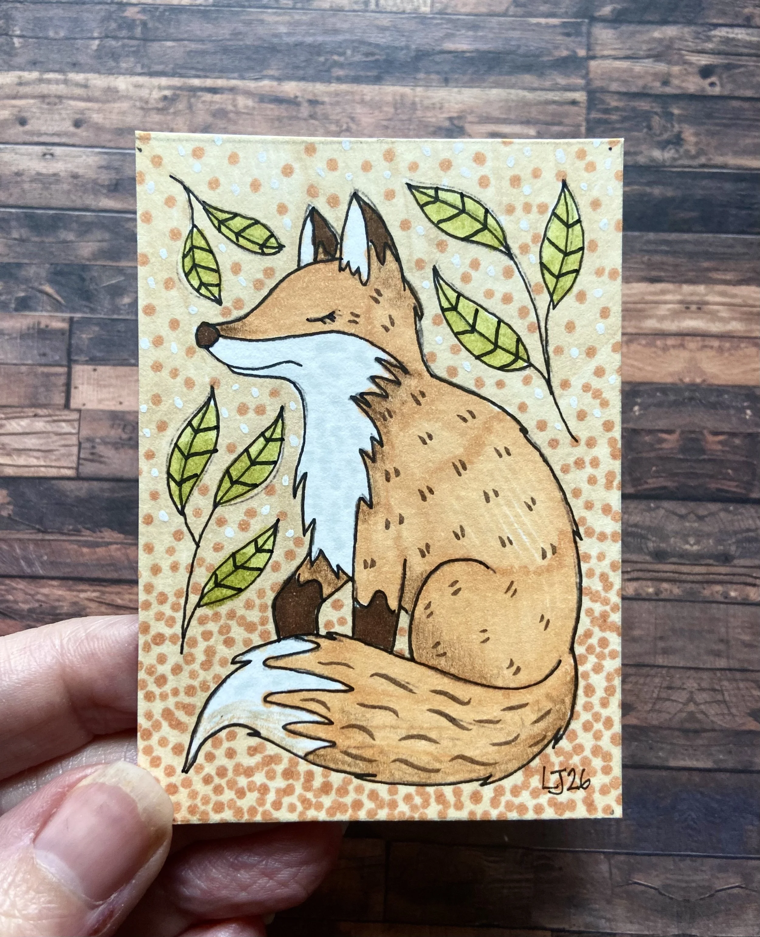Hand holding a card featuring a drawing of a fox with closed eyes, surrounded by green leaves on an orange dotted background.