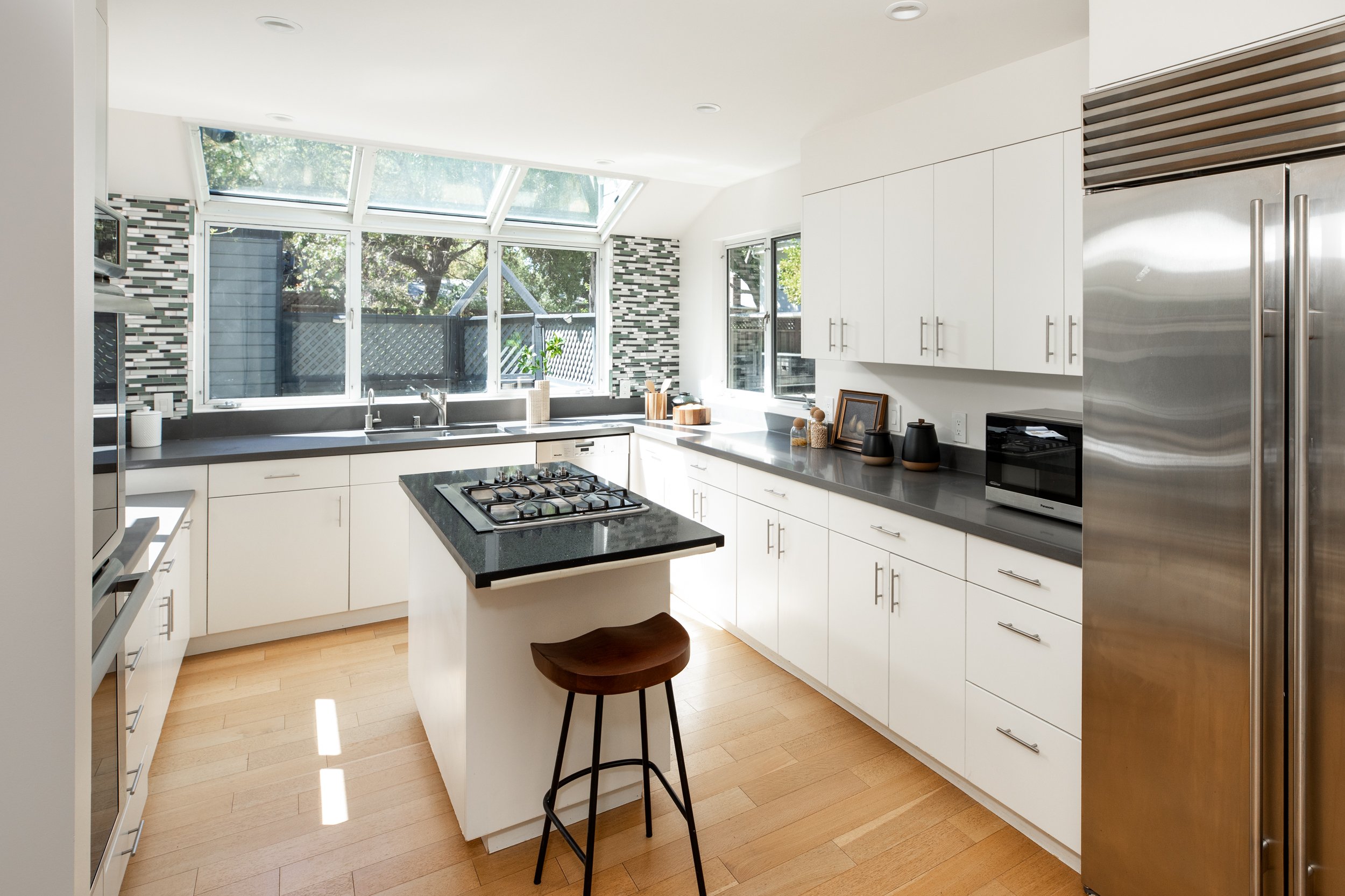 A modern kitchen with white cabinets, black countertops, a window, a small island with a wooden stool, and stainless steel appliances.