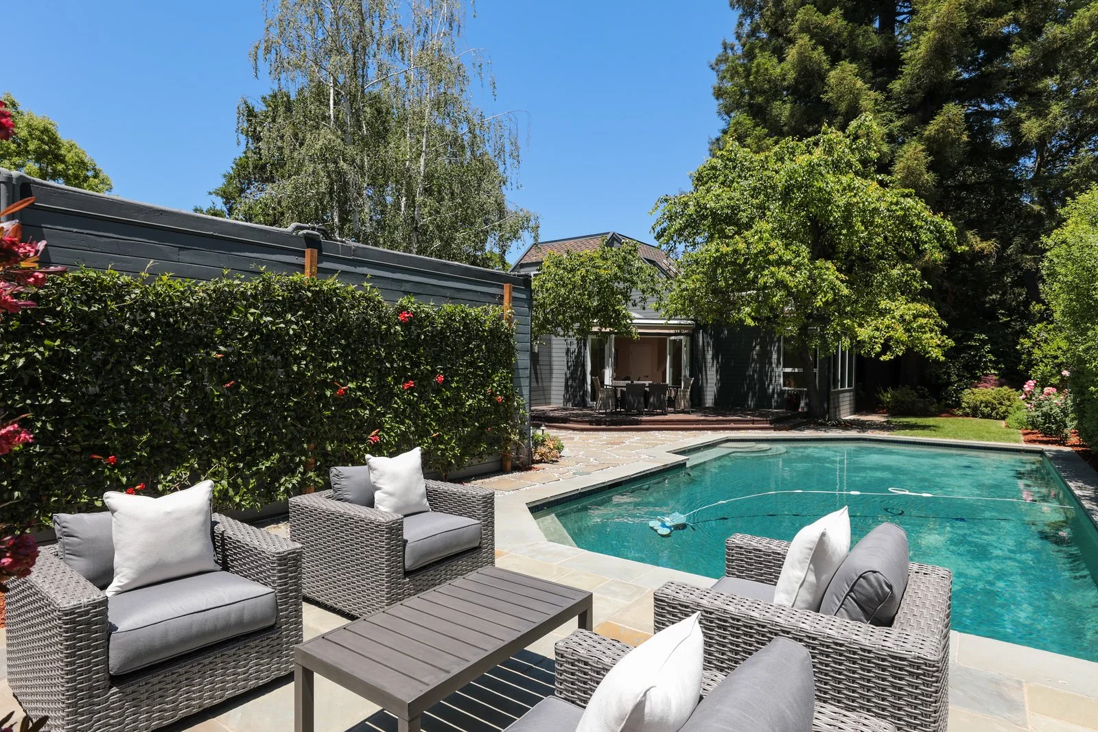 Outdoor backyard scene with a swimming pool, surrounded by wicker chairs with cushions, and a patio area with a dining table, framed by trees and a blue sky.
