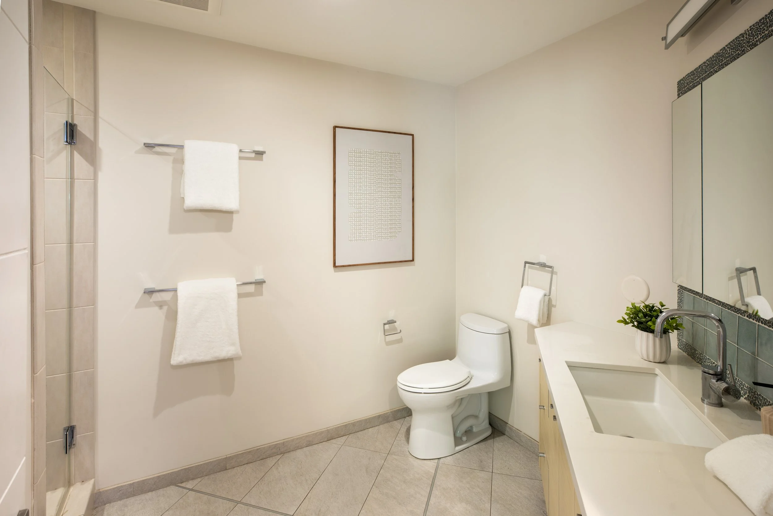 A modern bathroom with a toilet, sink, mirror, and towel racks with white towels, a framed abstract art piece on the wall, a small potted plant on the sink, and a glass-enclosed shower on the left.