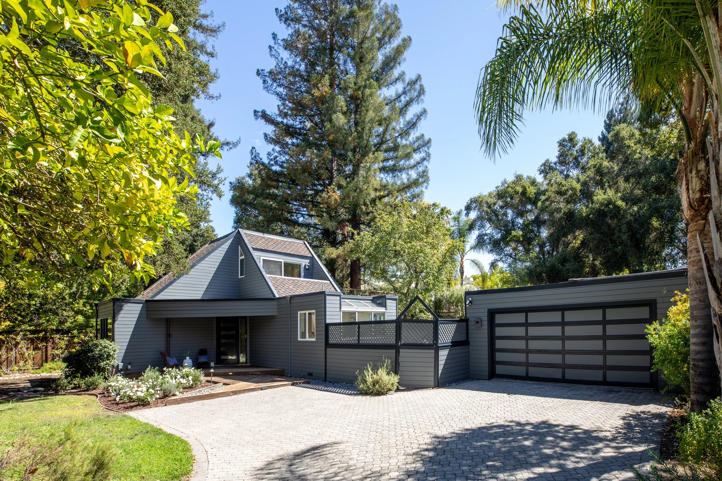 A modern gray house with a garage and large windows, surrounded by lush trees and a well-maintained garden with flowers, under a clear blue sky.