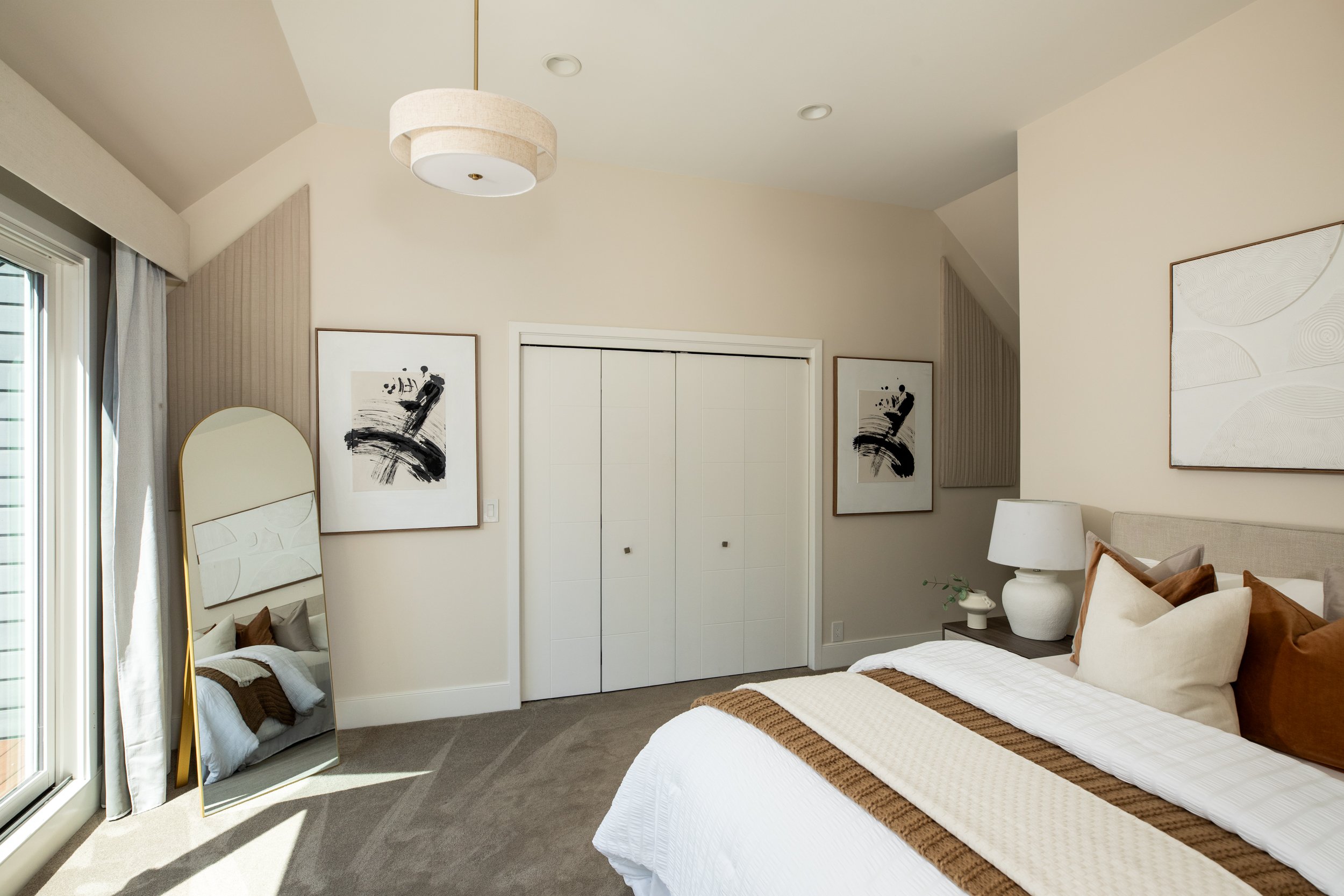 A modern bedroom featuring a large bed with white linens and brown accents, a standing mirror with a gold frame, abstract black and white artwork, a white closet with double doors, a window with light curtains, and a ceiling lamp.