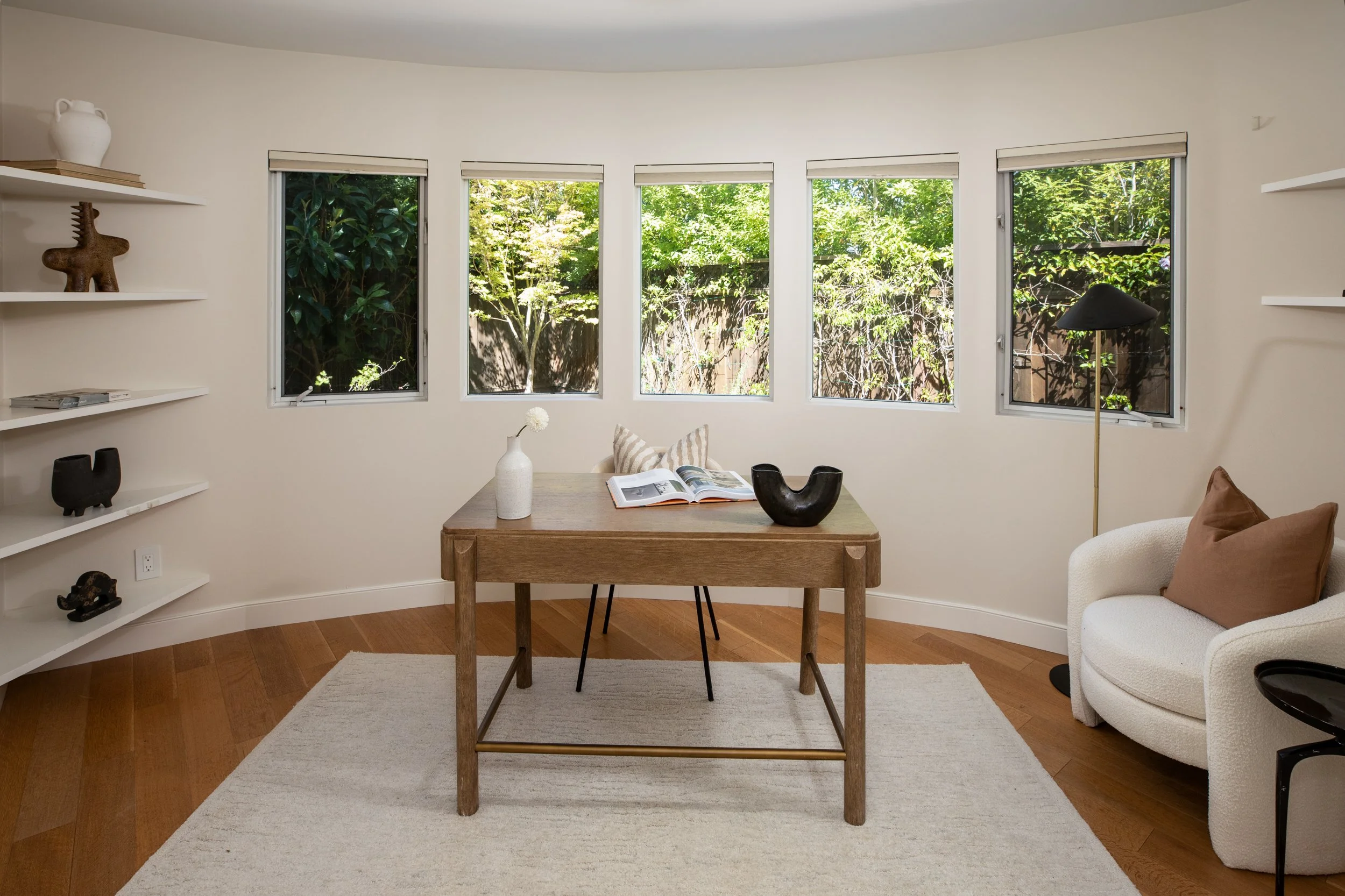 Living room with three large windows showing green trees outside, a small wooden dining table with open book and vases, a beige armchair with brown pillow, white built-in shelves with decorative items, a floor lamp, and a white rug on wooden flooring
