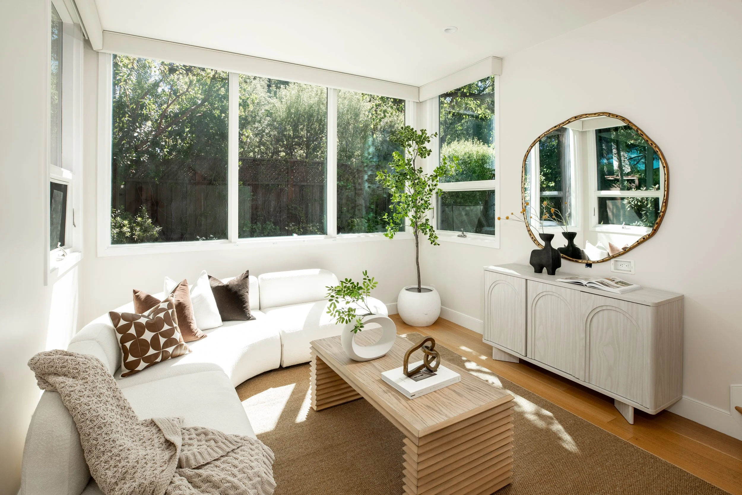 Bright living room with large windows showcasing greenery outside. Includes a white sectional sofa with brown and patterned pillows, a wooden coffee table, a potted plant, a white sideboard, and a decorative mirror on the wall.