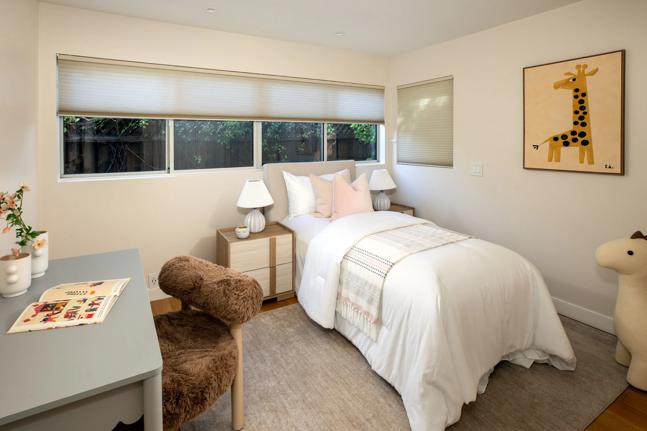 A cozy bedroom with a bed, nightstands, lamps, and a giraffe art on the wall, featuring a window with blinds and some greenery outside.