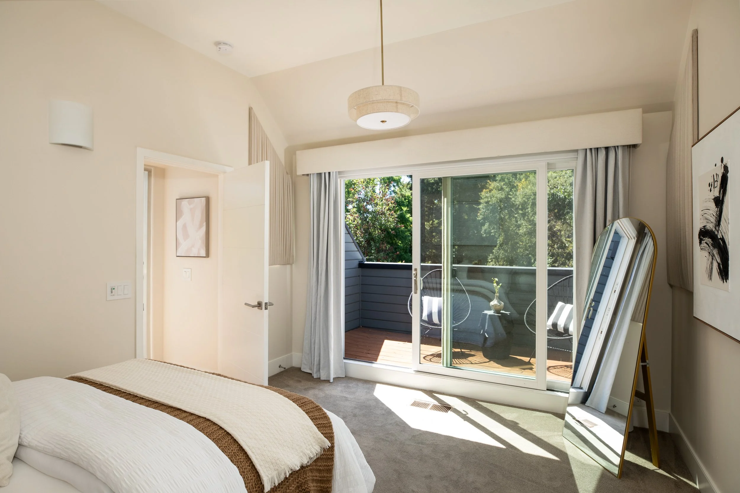 A bedroom with white walls, a bed with white bedding and a brown throw, a large sliding glass door leading to a balcony with outdoor chairs and a potted plant, and a full-length mirror leaning against the wall. Decor includes abstract art and wall-mo