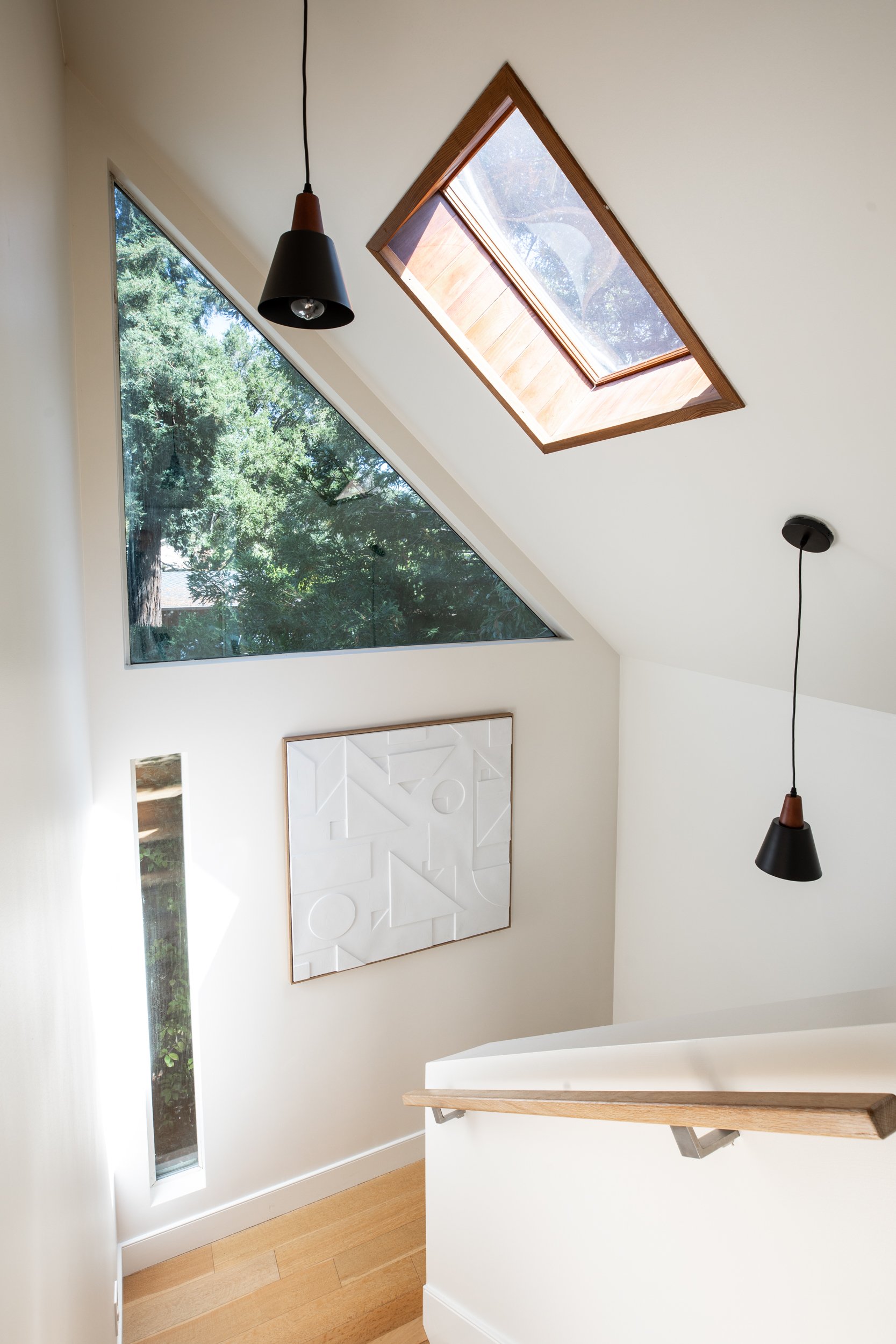 A modern staircase with a wooden handrail leading to a room with a large triangular window and a rectangular skylight in the ceiling, with a white wall art piece on the wall.