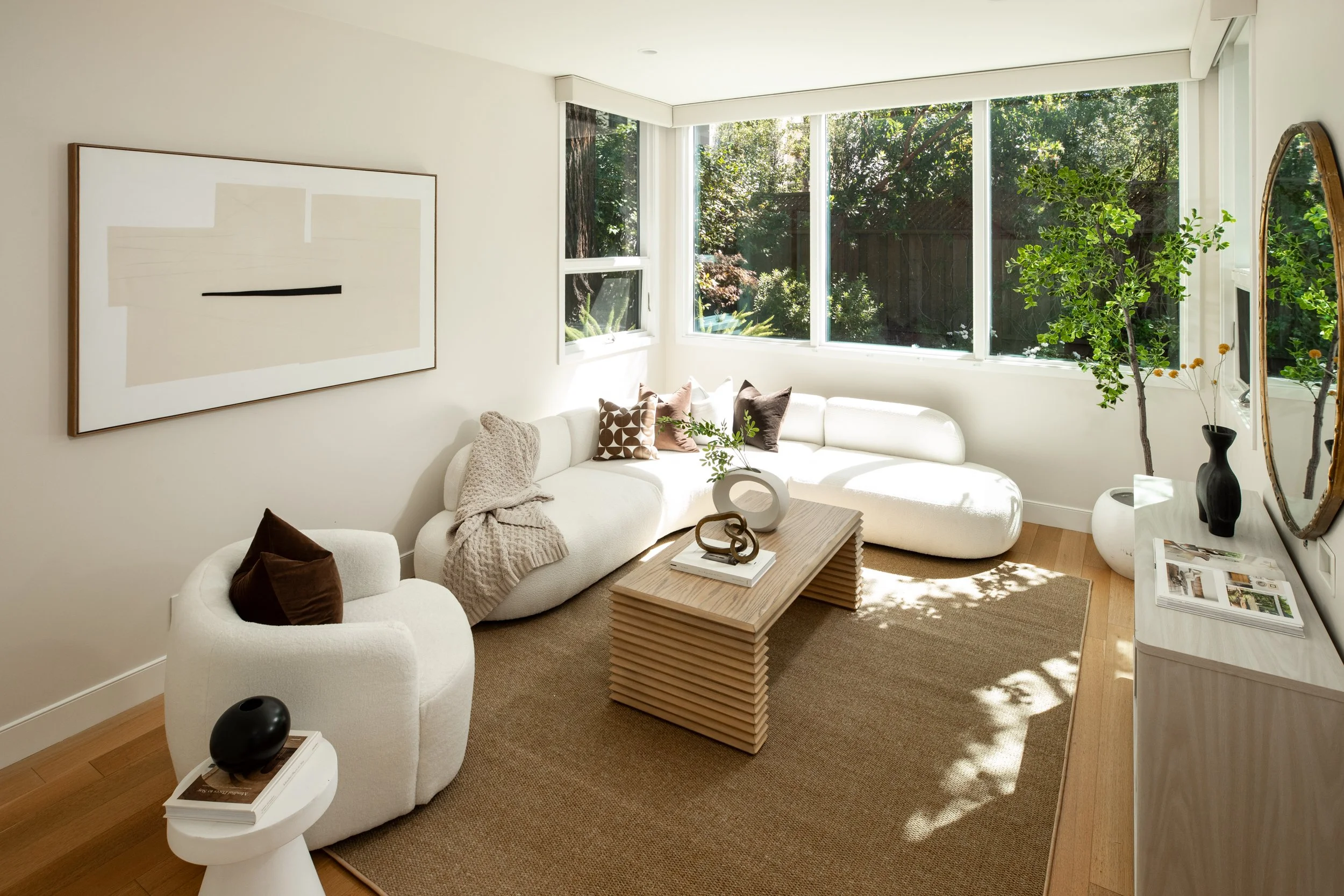 Bright living room with white curved sofa, decorative pillows, wooden coffee table, side table with books and black spherical decor, large windows showing green trees outside, artwork on wall, potted plant, and mirror reflecting interior.