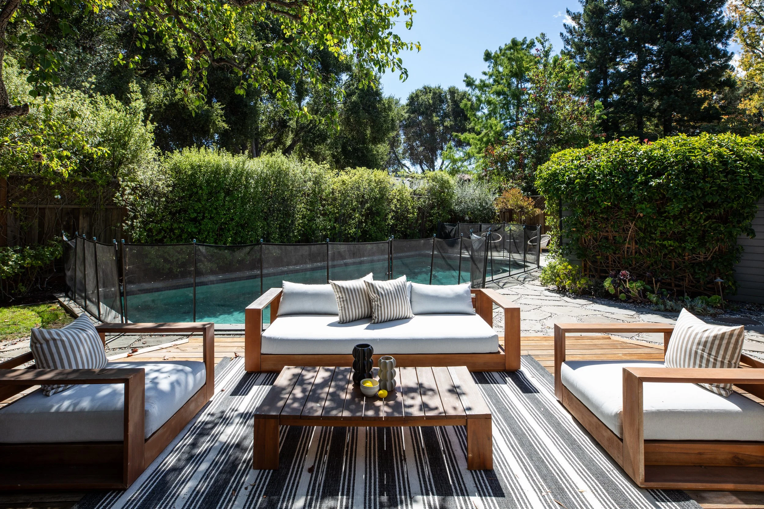 A cozy outdoor backyard patio with wooden lounge furniture, striped cushions, and a striped rug, surrounded by lush green trees and bushes, and a swimming pool with a safety net.