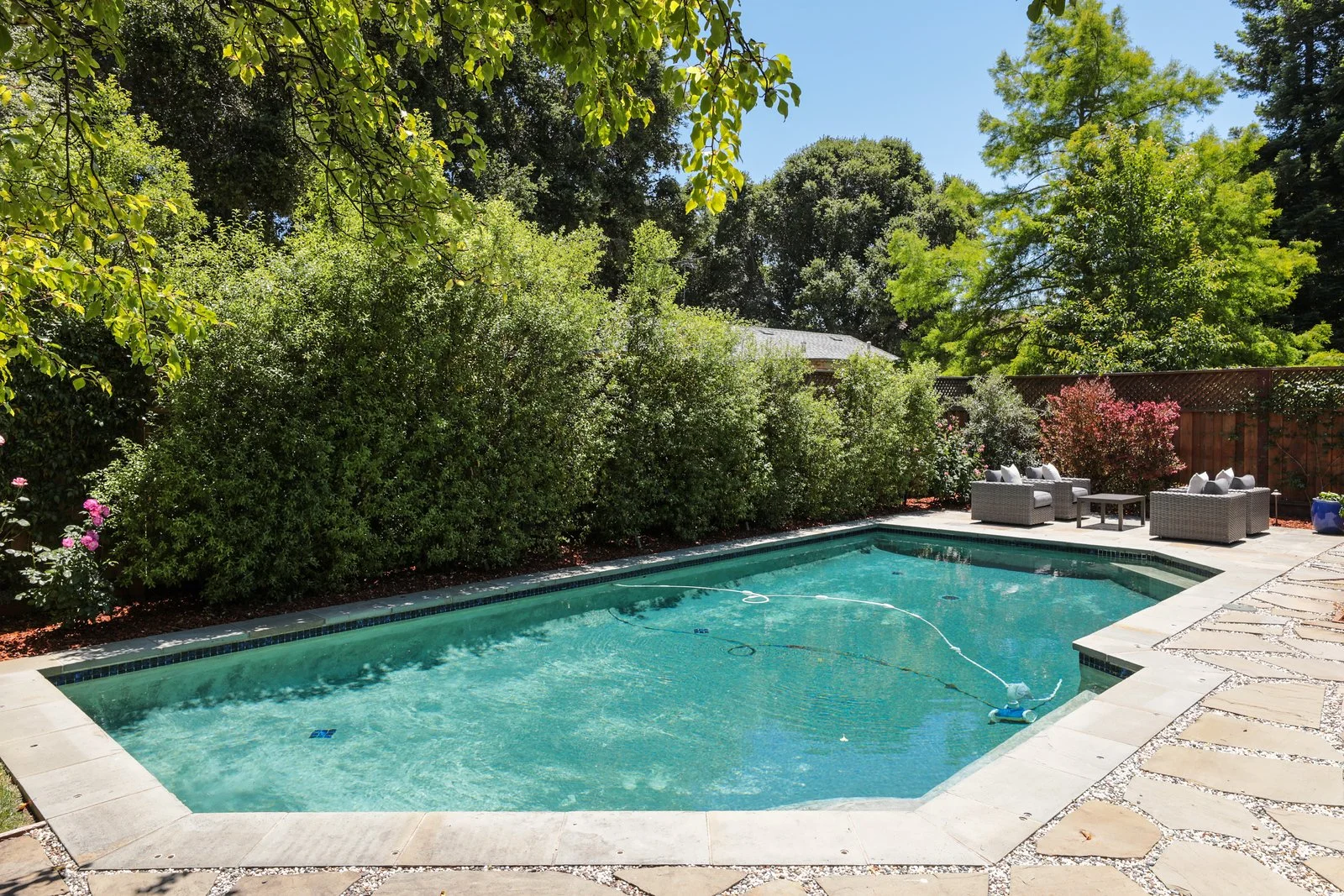Backyard swimming pool with outdoor seating area, surrounded by lush green bushes and trees, during sunny day.