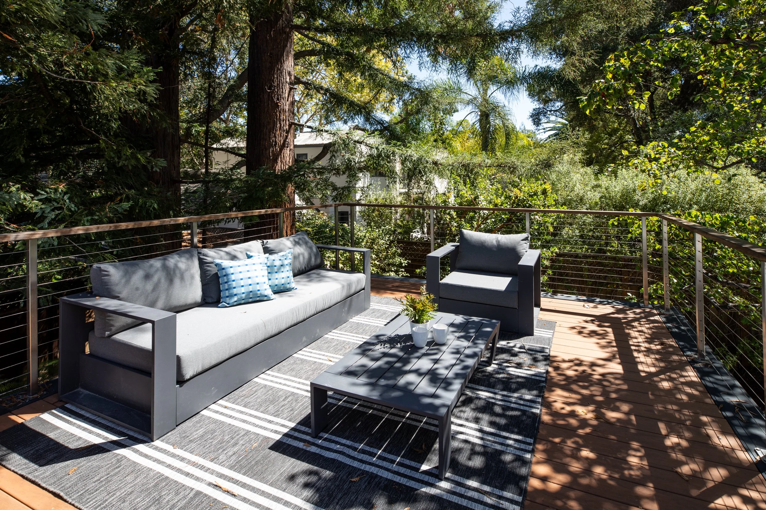An outdoor deck with a gray sofa and matching armchair, decorated with blue and white cushions, a striped rug, and a small table with white vases, surrounded by lush green trees and a metal railing.