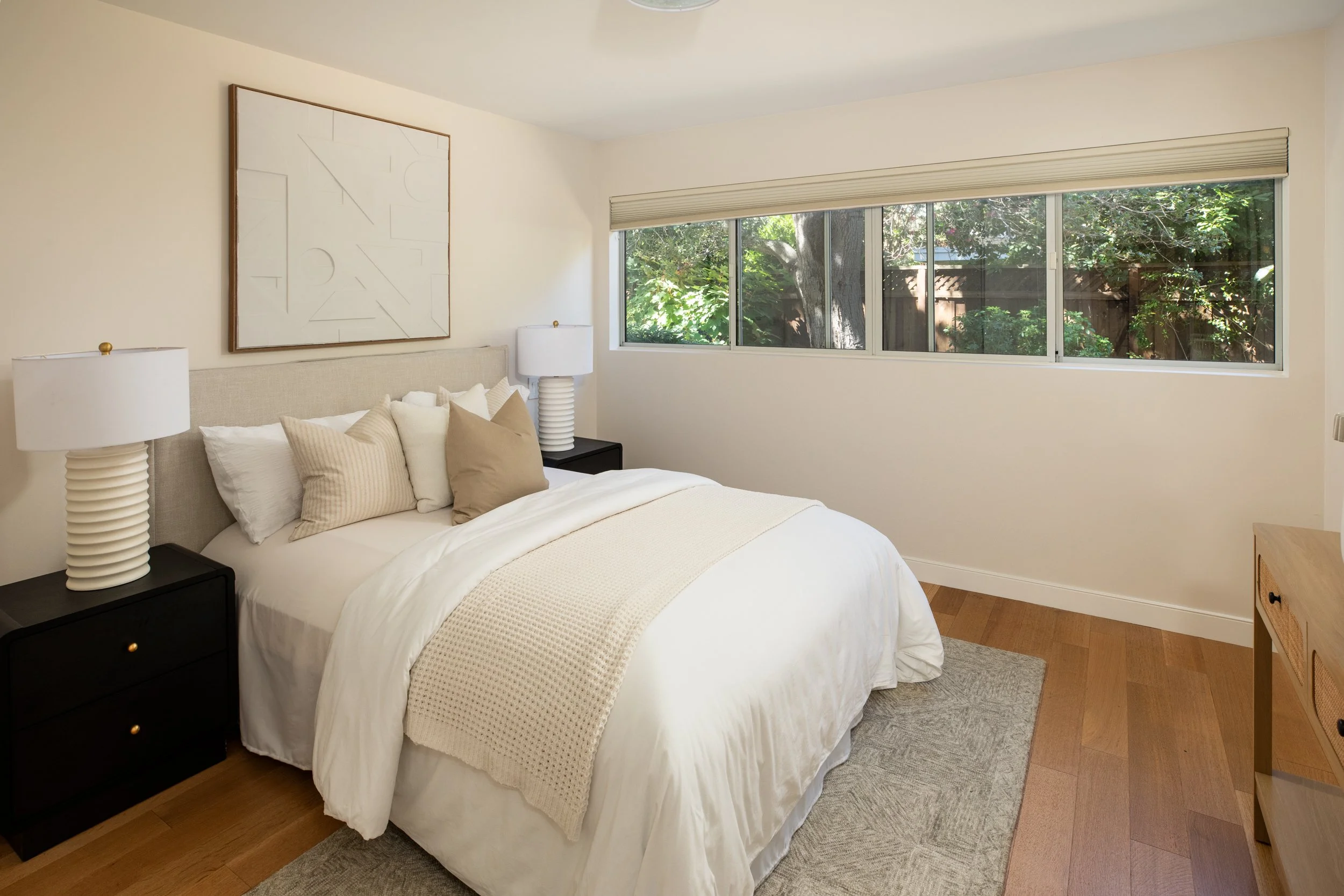 A cozy bedroom with a bed dressed in white and beige pillows, black nightstands with white lamps, a large window with a view of trees and nature, a framed abstract wall art, a beige rug, and wooden flooring.