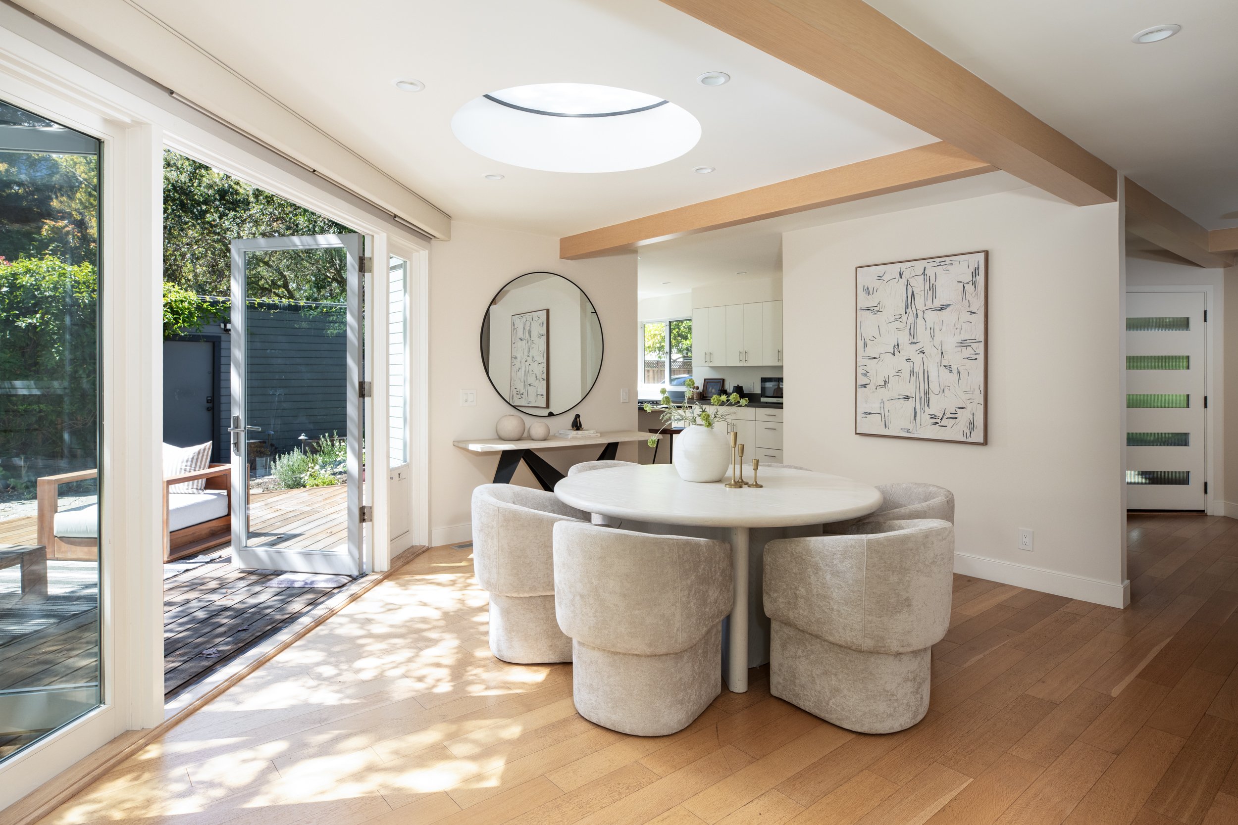 A bright, modern living room with sliding glass doors opening to an outdoor patio, a round dining table with beige upholstered chairs, a wall mirror, and white cabinets in the kitchen area.