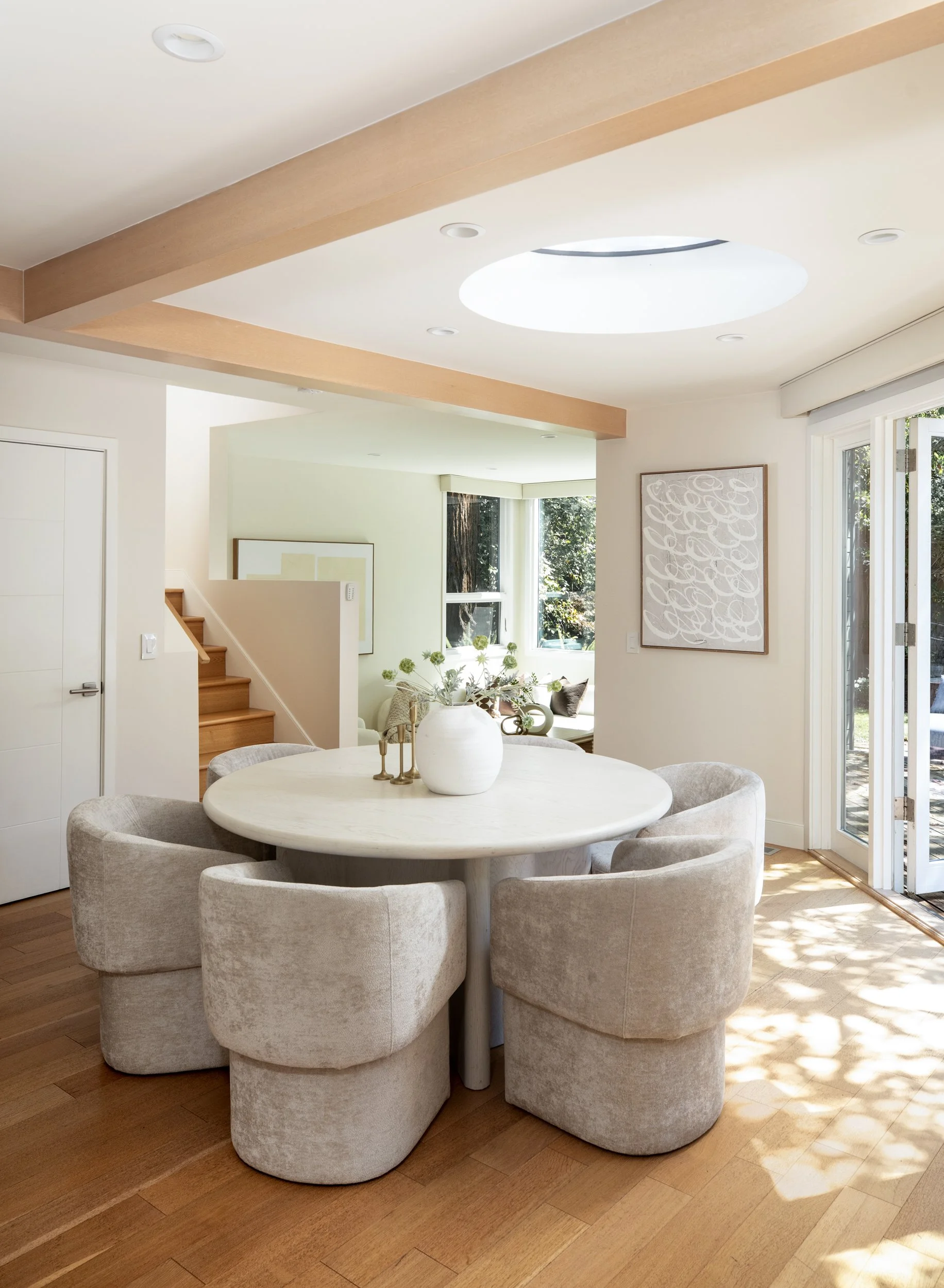A round table with six beige upholstered chairs, a white vase with flowers, set in a bright living space with large windows and wood flooring.