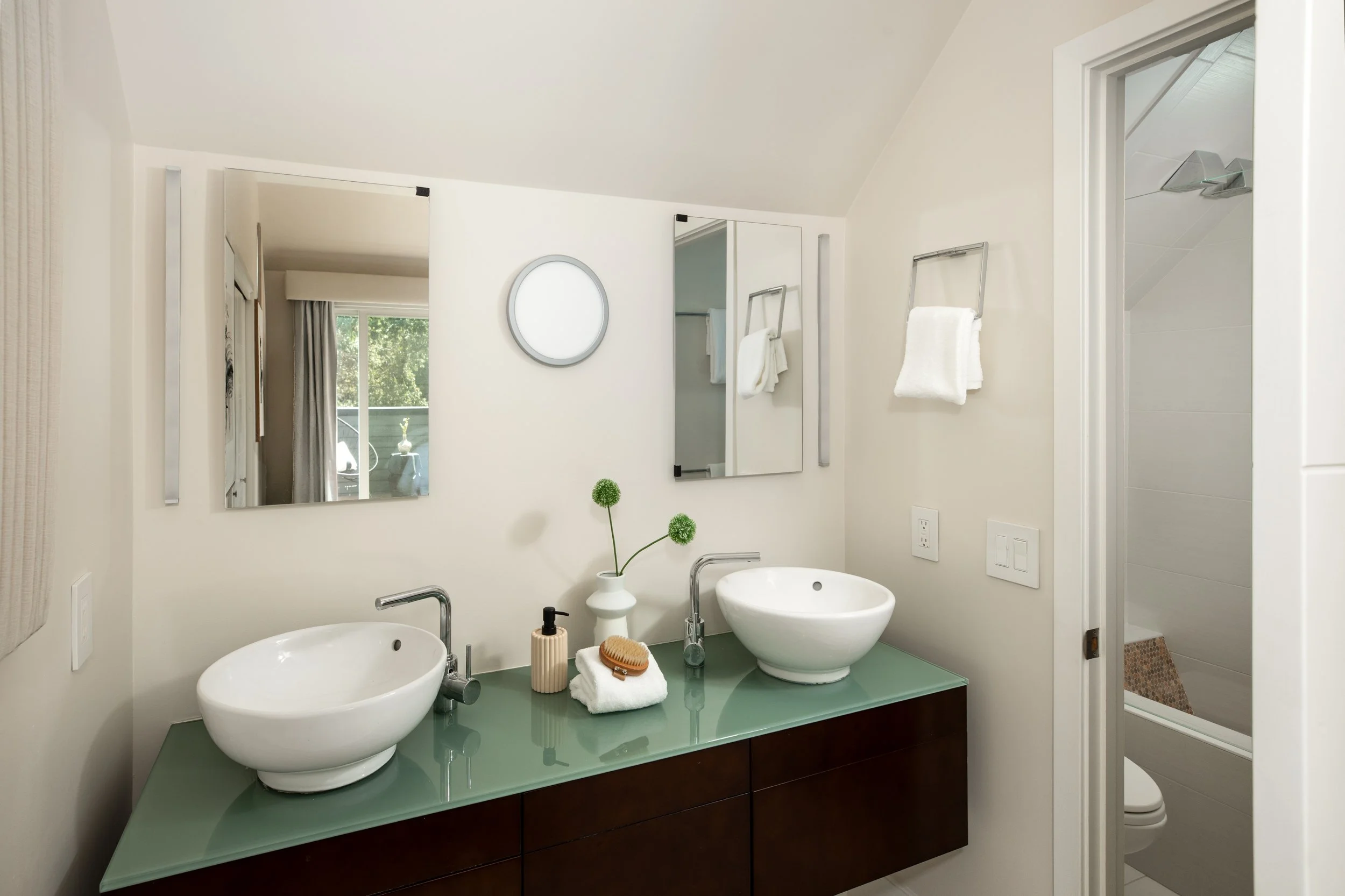 Modern bathroom with two white vessel sinks on a green countertop, mirrors on the wall, towels hanging, a window with curtains, and decorative items including a plant and toiletries.