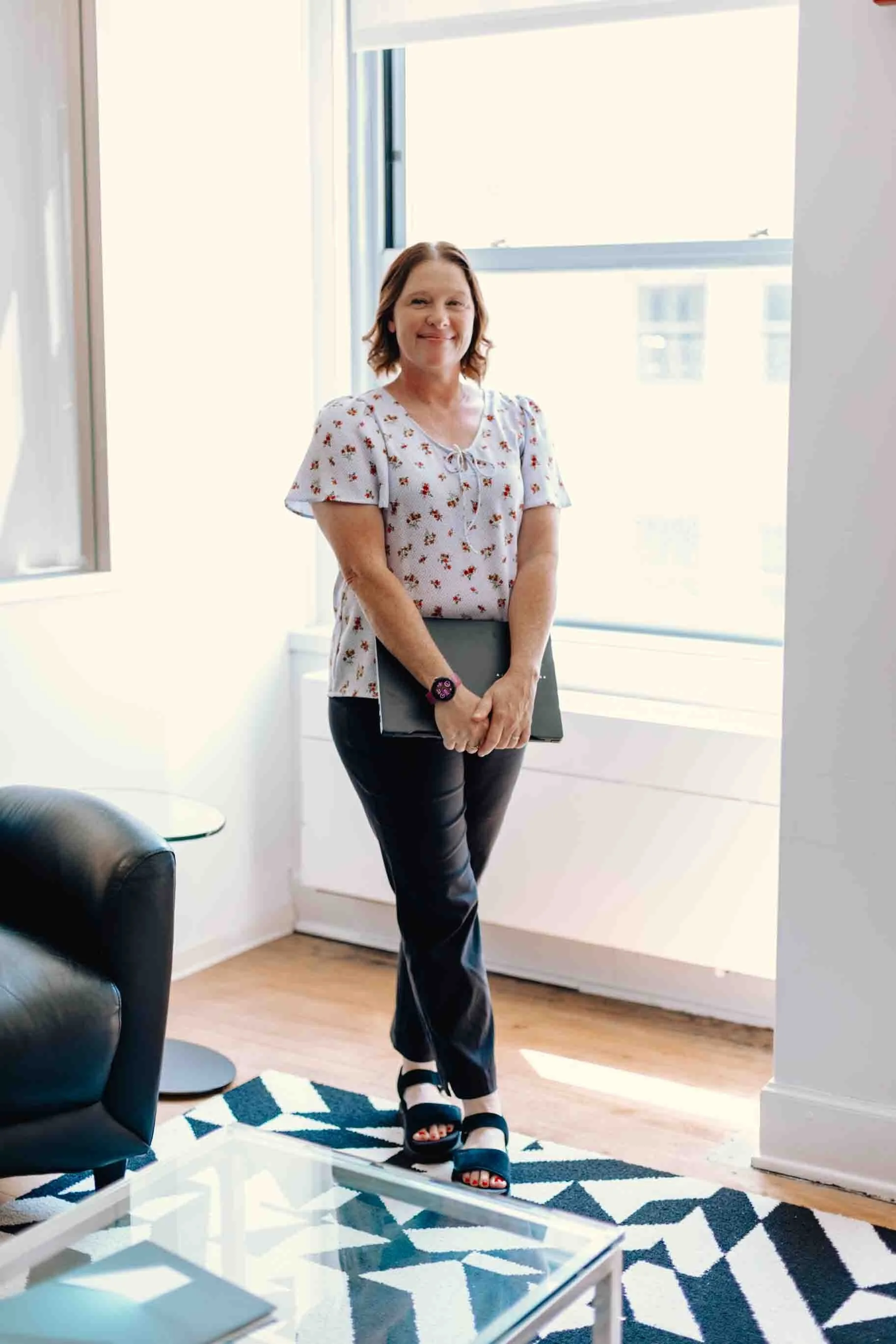 Liz Chapman, podcast strategist, is standing by a window smiling and has her laptop in her hands.